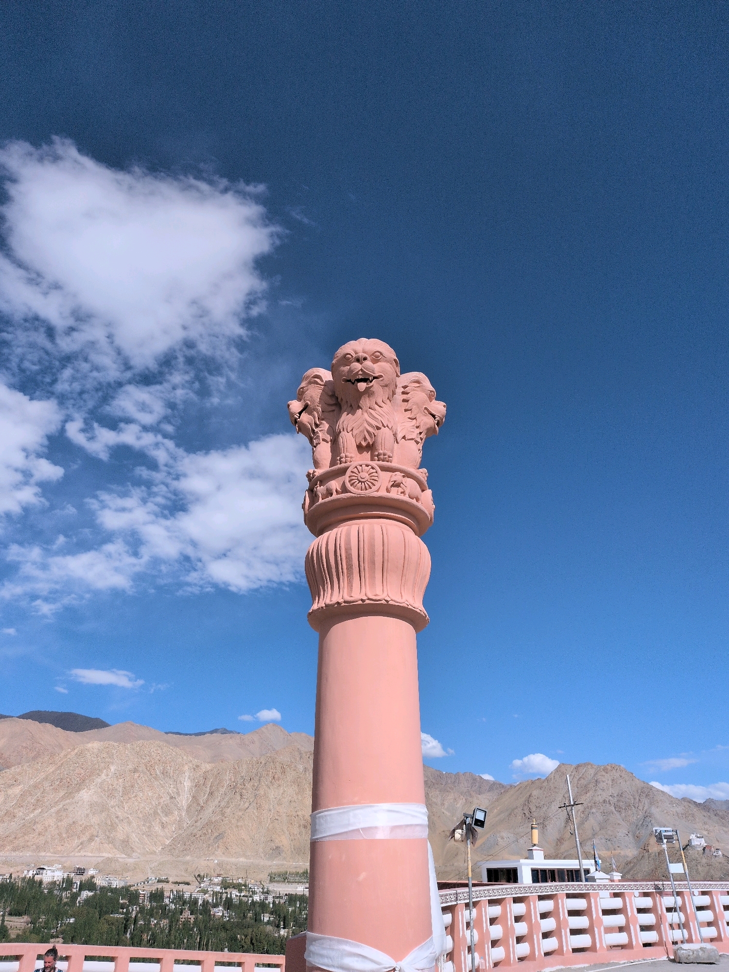 A low-angle, medium shot captures the Ashoka pillar, a prominent monument, set against a vibrant blue sky with scattered white clouds. The pillar, rendered in a soft pink hue, features a sculpture of four lions at its apex, with one lion prominently displayed in the foreground, its mouth open. Below the lions, a wheel with eight spokes is visible. The pillar itself is cylindrical with a fluted texture at the top, and a white band wraps around its lower section.
In the background, a town is nestled at the base of rugged, tan-colored mountains. The town is characterized by a dense arrangement of buildings and numerous green trees. A pink railing with white horizontal bars runs along the foreground, suggesting an elevated viewing platform. A white building with a dark roof is visible beyond the railing, and a single street lamp on a pole stands near it. The overall environment suggests an arid, mountainous region with a clear, sunny day.