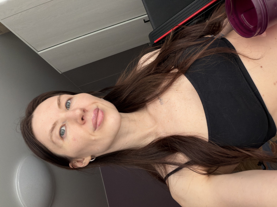 A woman with long, brown hair, wearing a black ribbed top, is captured in a selfie. She has fair skin, blue eyes, and a slight smile. Her hair is parted to the side and cascades around her shoulders. A dark purple shaker bottle is visible in the upper right corner. The background includes a white cabinet or wall, a black and red piece of equipment, and a gray wall. The image has a slightly rotated perspective.