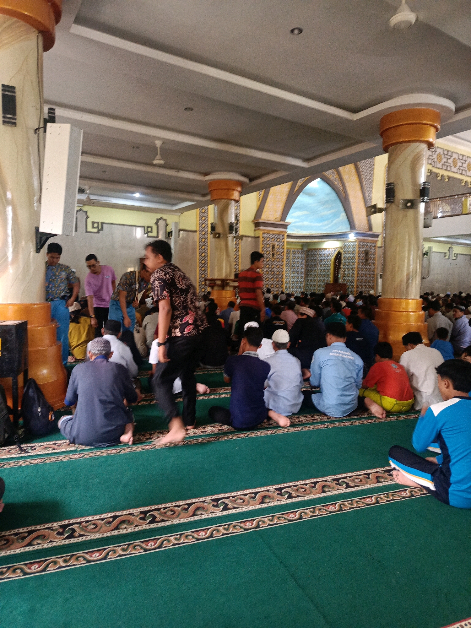 The image depicts a scene inside a mosque in Magetan, Indonesia, during what appears to be a gathering or prayer service. A large number of men are seated on a green carpet with ornate patterns, facing towards the front of the mosque. Many are wearing traditional Islamic attire, such as skullcaps and sarongs, while others are in casual clothing.
In the foreground, a man in a dark blue outfit is seated facing away from the camera. To his right, a person in a bright blue shirt and dark pants is also seated. Further to the right, a person in a red shirt and yellow pants is visible. The carpet is a prominent feature, with intricate designs in brown, gold, and black.
The mosque's interior features large, decorative pillars with gold accents, supporting a high ceiling with intricate molding. The walls are adorned with patterned tiles and a large arched decoration that resembles a sky with clouds. Several ceiling fans are visible, suggesting a warm climate.
In the background, more people are seated, and some are standing. Two men in patterned shirts are standing near the left side of the image, next to a large orange structure that appears to be part of the mosque's architecture or possibly large speaker cabinets. One man in a striped shirt is standing further back, closer to the arched decoration. The overall atmosphere is one of communal gathering and devotion.