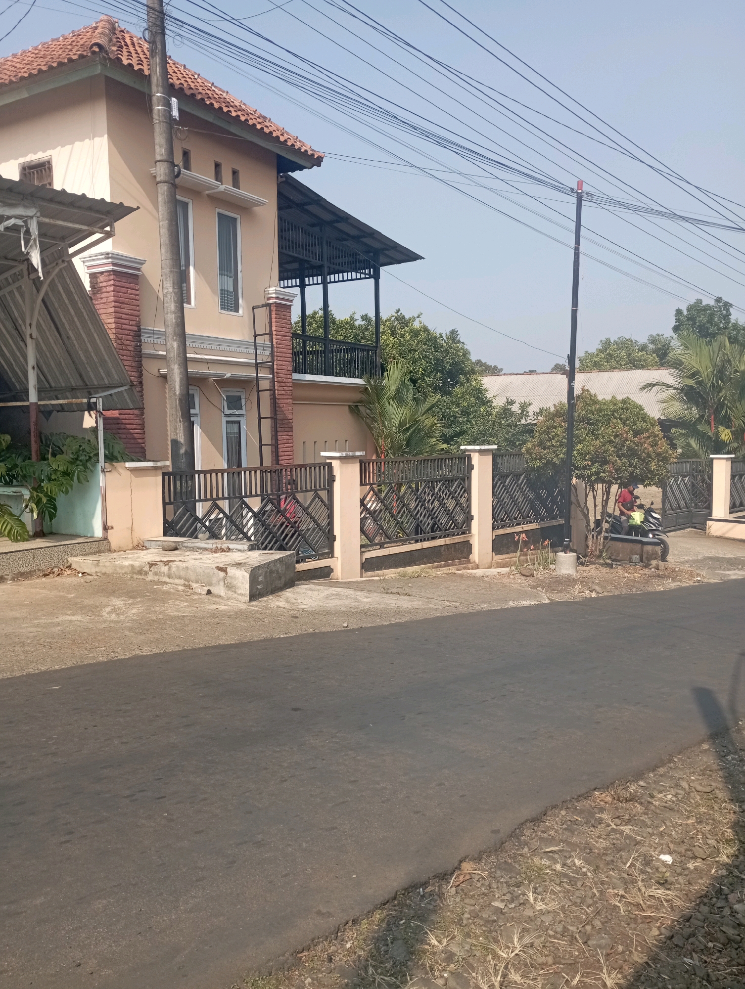 In the city of Pendem, Indonesia, a residential scene unfolds. The primary subject is a two-story house, painted in a neutral beige tone, with a terracotta-tiled roof. Red brick accents adorn the base of some pillars and parts of the facade. A black metal fence with a diamond pattern encloses the property, with beige concrete pillars supporting it. A motorcycle, with a rider wearing a red shirt and a dark helmet, is parked near the fence on the right side of the image. The house has several windows, some with white frames and dark shutters, and a black metal balcony railing on the upper floor. A metal ladder is attached to the exterior wall of the house, leading to the upper level. A gray utility pole stands prominently in front of the house, with numerous electrical wires extending across the clear blue sky. The foreground features an asphalt road. Vegetation, including trees and plants, surrounds the house and fence, with some greenery visible to the left, behind the fence, and on the right. The ground near the road and fence is covered with dirt, small stones, and dry grass. The lighting suggests it is daytime, with the sun casting shadows.