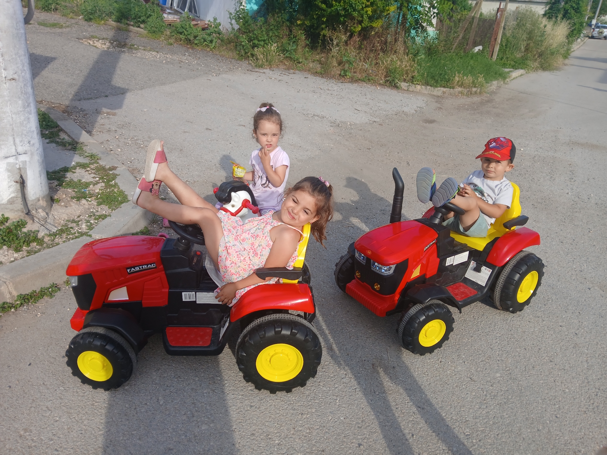 Three young children are playing outside in the daytime. Two girls and a boy are seated on red toy tractors. The girl in the foreground is sitting on the left tractor, leaning back with her legs extended. She is wearing a floral print dress and pink sandals. Her expression is happy and playful.
Behind her, another girl is sitting on a black and red toy tractor. She is wearing a light purple t-shirt and has a serious expression. She is holding a yellow and black object, possibly a snack.
On the right tractor sits a boy wearing a grey t-shirt, green shorts, and a red baseball cap with a Spider-Man logo. He is looking forward with a neutral expression. The tractors are red with black accents and yellow wheels.
The scene appears to be on a paved road or driveway in a residential area. There are some overgrown plants and trees in the background, suggesting a rural or semi-rural setting. The lighting indicates it is daytime, possibly late afternoon due to the shadows. The children's relaxed postures and smiles suggest a casual and fun activity. Visible text on the left tractor reads "FASTRAC".