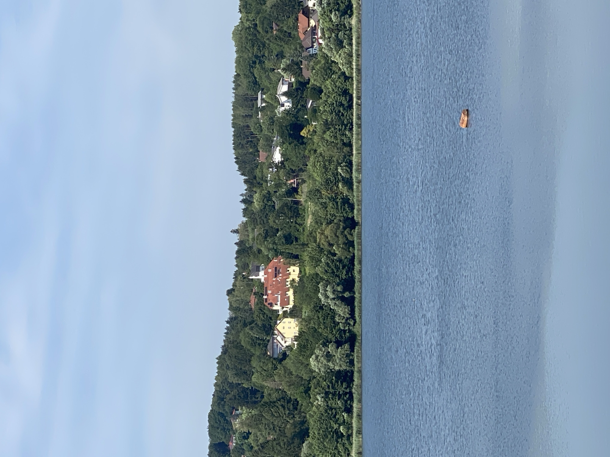 The image shows a wide body of blue water in the foreground, likely a lake, with ripples on its surface. In the middle ground, a treed hillside is visible, dotted with several houses and a prominent building with a red-tiled roof. A church spire can be seen above the trees. The background consists of a hazy blue sky. A small, light brown object, possibly a piece of driftwood, floats in the water towards the right side of the frame. The scene appears to be a scenic view of a lakeside town, likely Purkersdorf, Austria.