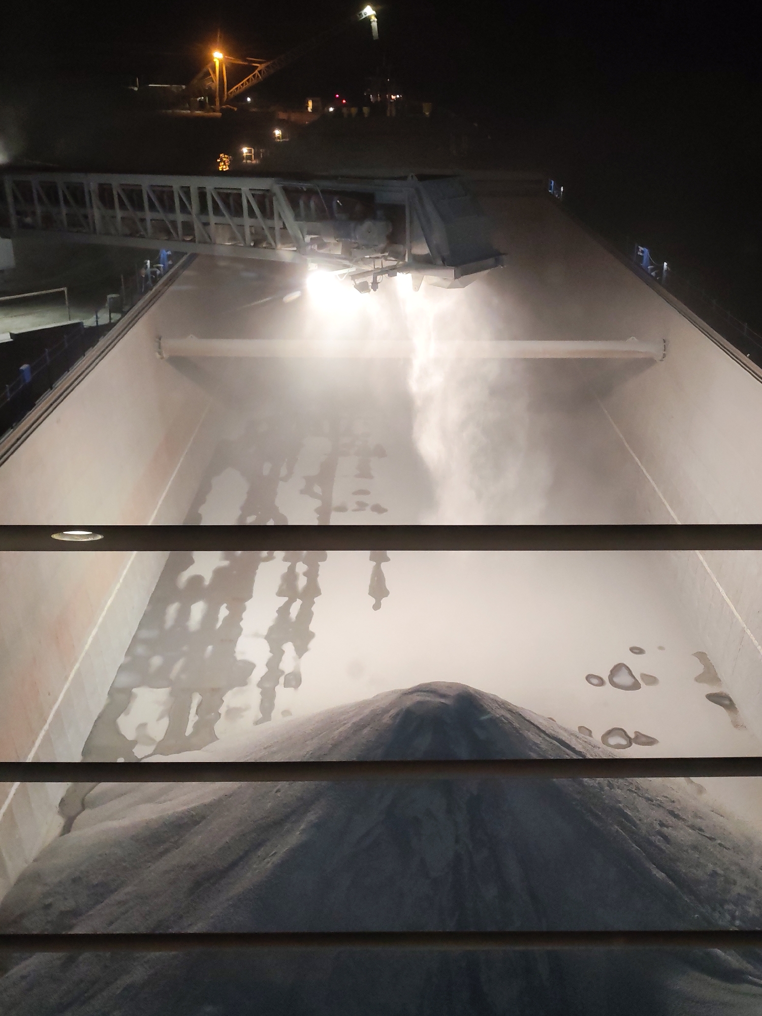 The image depicts the interior of a ship's cargo hold being filled with a large mound of gray material, possibly granular or powdered, under bright artificial lighting. The loading equipment, including a conveyor system, is visible above. The scene is likely set at night, given the surrounding darkness and the prominence of artificial lights. No people are directly visible inside the hold, although there might be shadows suggestive of their presence. The image is framed by what appears to be a set of horizontal railings, suggesting the viewpoint is from an elevated position above the cargo hold. Without additional information, a definitive determination of the location, including Shcheleiki, Russia, is not possible based solely on this image.