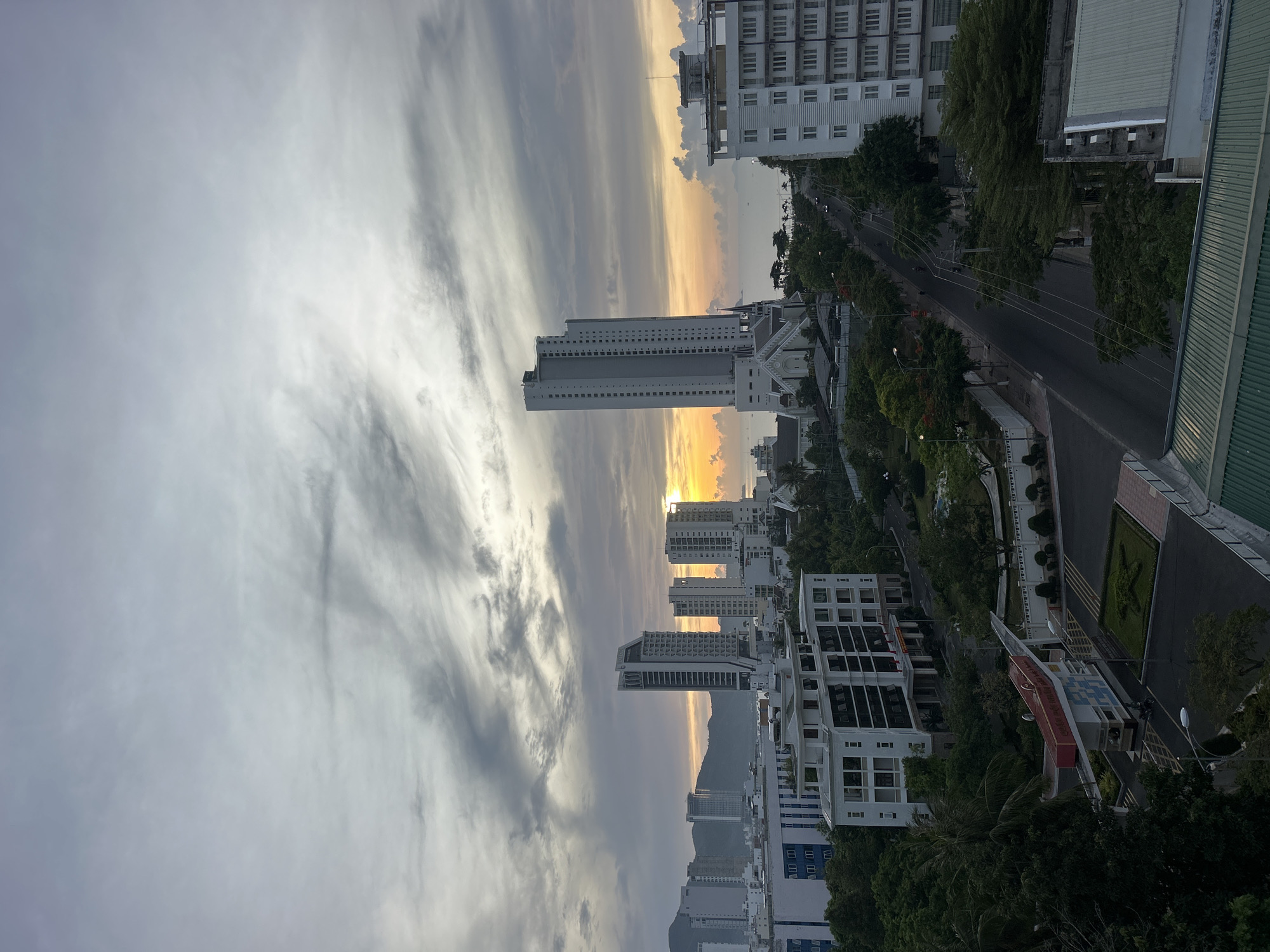 The image is a view over Nha Trang, Vietnam, likely from an elevated position. It shows multiple high-rise buildings, some appearing residential or hotel-style. The sky is filled with stratus clouds that are striated. A road runs through the scene, and trees are abundant along the side of the road. The sun appears to be setting on the horizon. Some buildings are multi-story and light-colored. The lighting in the image suggests either sunrise or sunset, with some color in the sky. The buildings feature different architectural styles.