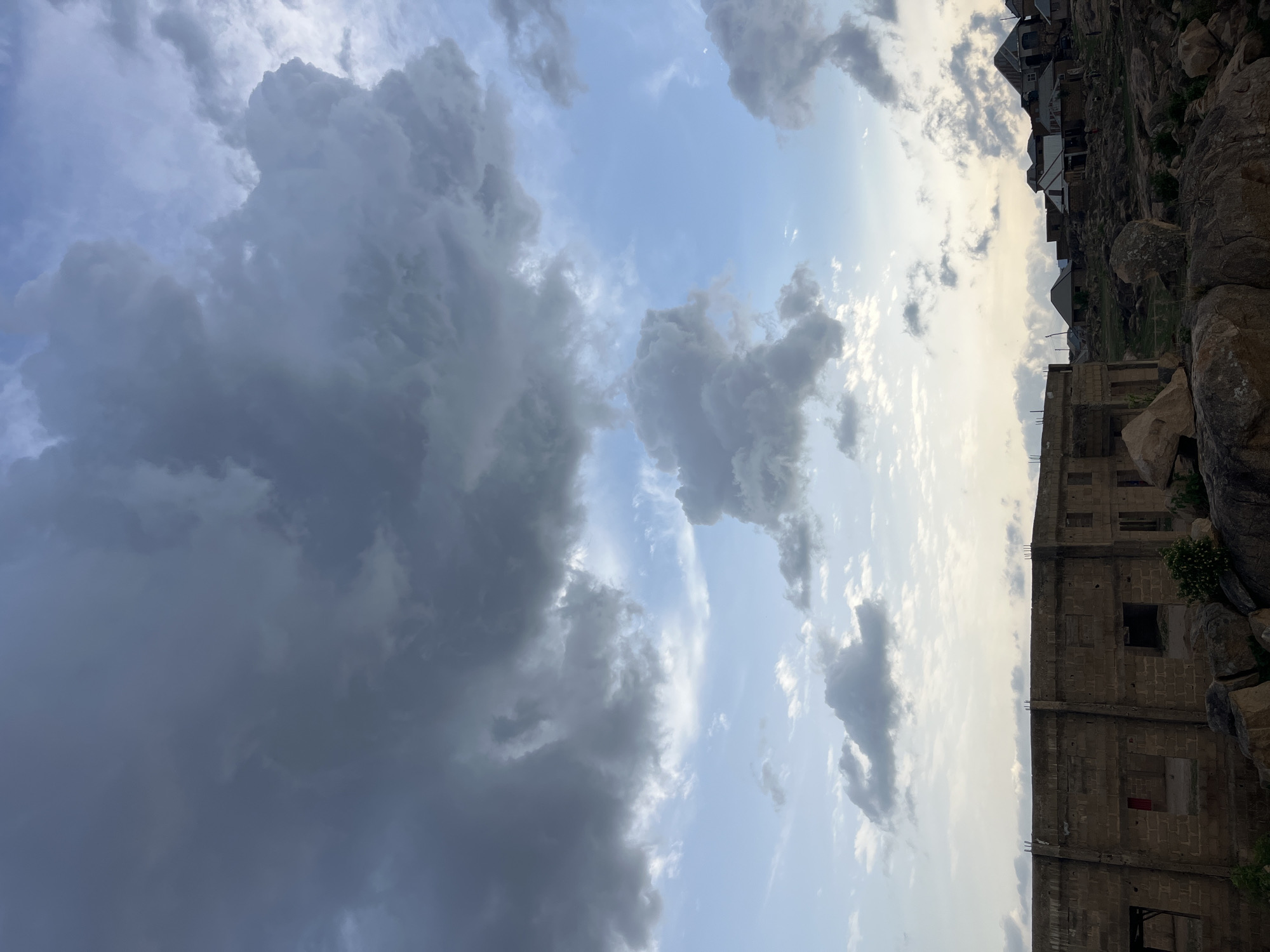 The image depicts a partly cloudy sky over Jos, Nigeria. The left side of the image shows thick, dark storm clouds, while the right side has lighter, more scattered clouds with glimpses of blue sky. The foreground features a grey, unfinished concrete building with empty window openings. Adjacent to the building are large rocks and sparse vegetation. In the distance, atop a rocky hill, a cluster of houses is visible.