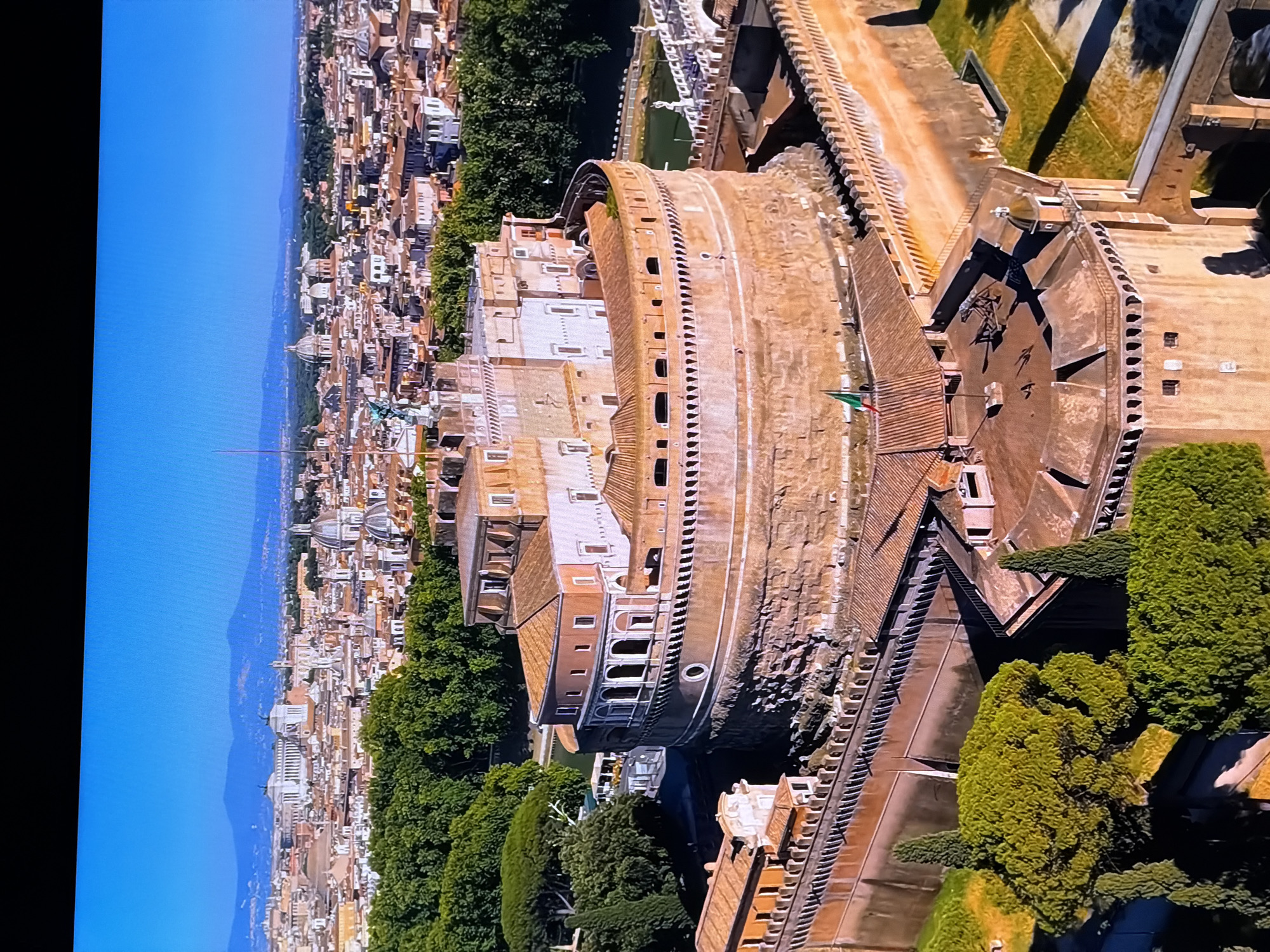 This is an aerial view of Castel Sant'Angelo in Rome, Italy. The cylindrical fortress, constructed of brick and stone, is a prominent structure in the foreground, with its crenellated walls and tiered architecture. Red-tiled roofs of surrounding buildings are visible, along with dense green trees and a body of water, likely the Tiber River. In the background, the cityscape of Rome extends to the horizon, featuring numerous domed buildings and distant hills under a clear blue sky. The image does not show the city of Chirchik, Uzbekistan.