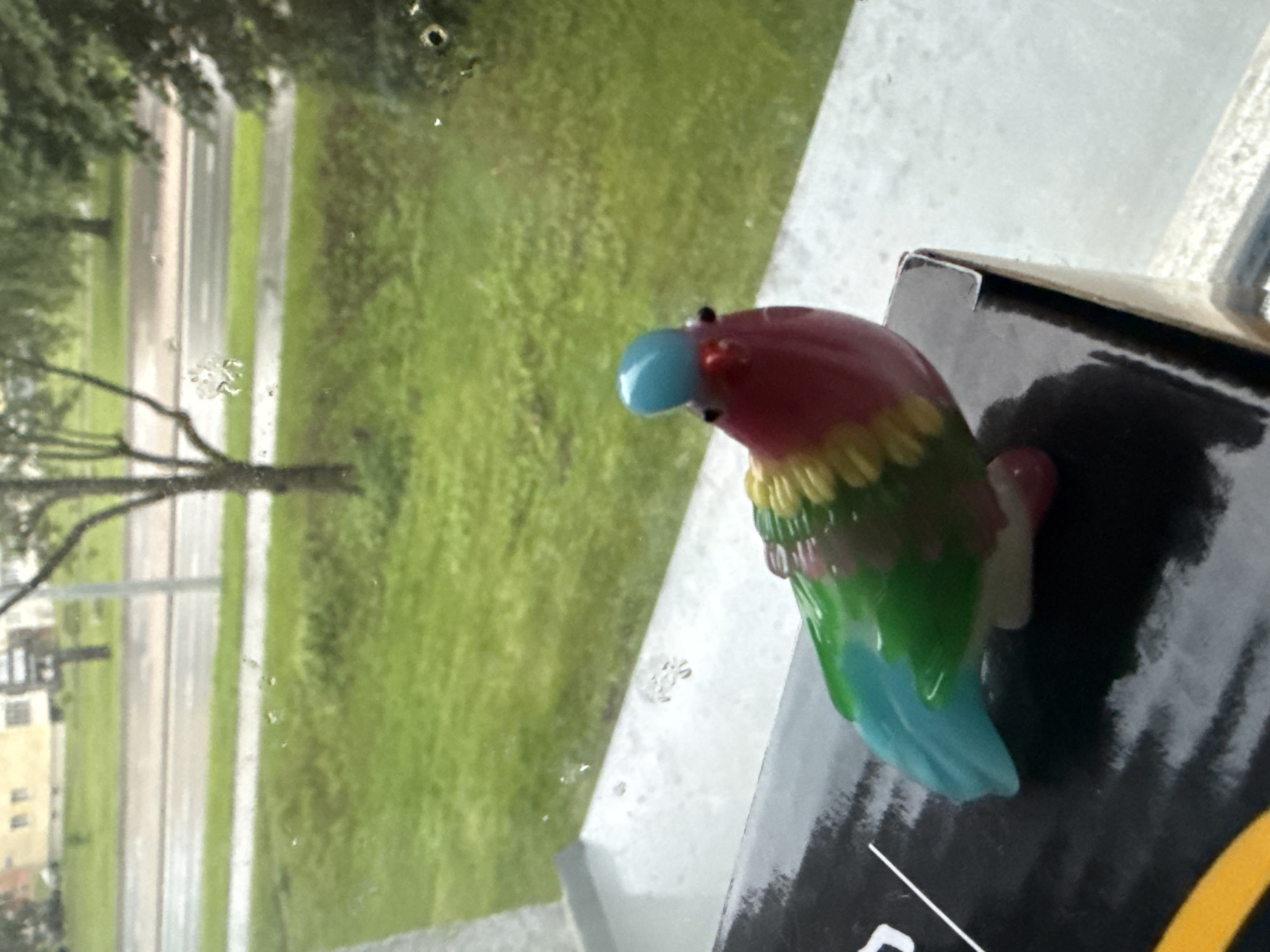 A close-up shot captures a colorful, small plastic bird figurine positioned on the edge of a black box. The figurine, predominantly red with blue, green, and yellow accents, has a bright blue crest. Its eyes are small and black, and its beak is red. The background is a blurred view through a window, showing a grassy area, trees, and a street with buildings in the distance. Raindrops are visible on the windowpane, suggesting inclement weather. The scene is likely set in a residential area of Tallinn, Estonia, given the architectural style of the distant buildings and the typical urban greenery. The overall impression is one of an indoor object placed near a window with an outdoor view.