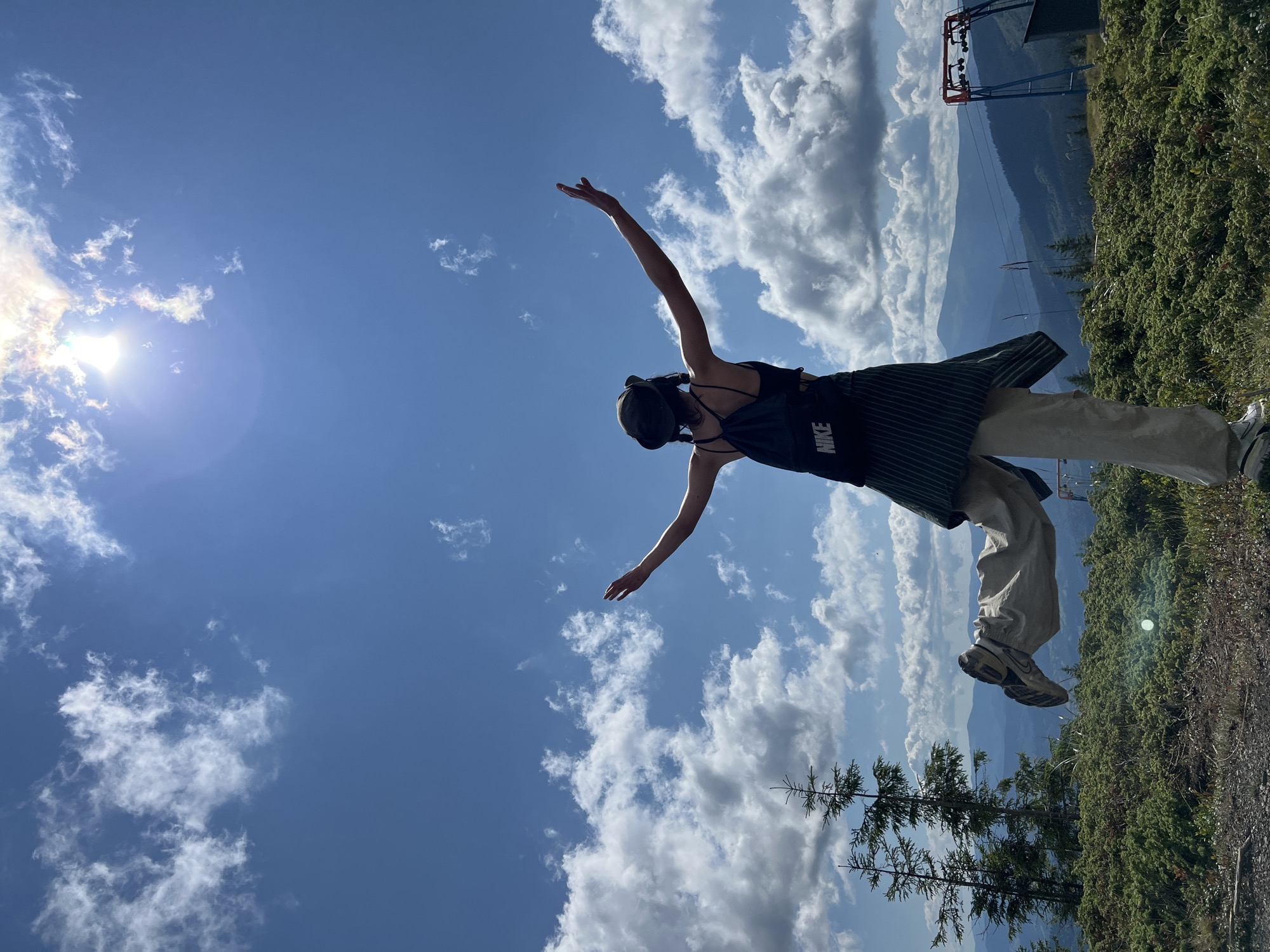 The image shows a person standing on a grassy hillside with their arms outstretched towards the sun. The person is wearing a hat, a black tank top, and light-colored pants. They have a black backpack with the word "NIKE" visible. The background features a vast sky with white clouds and a bright sun. In the distance, there are rolling hills and mountains, suggesting a mountainous region. There are also some evergreen trees on the left side of the frame and what appears to be part of a ski lift structure on the right. The overall scene conveys a sense of freedom and connection with nature. The location is identified as Chorna Tysa, Ukraine.