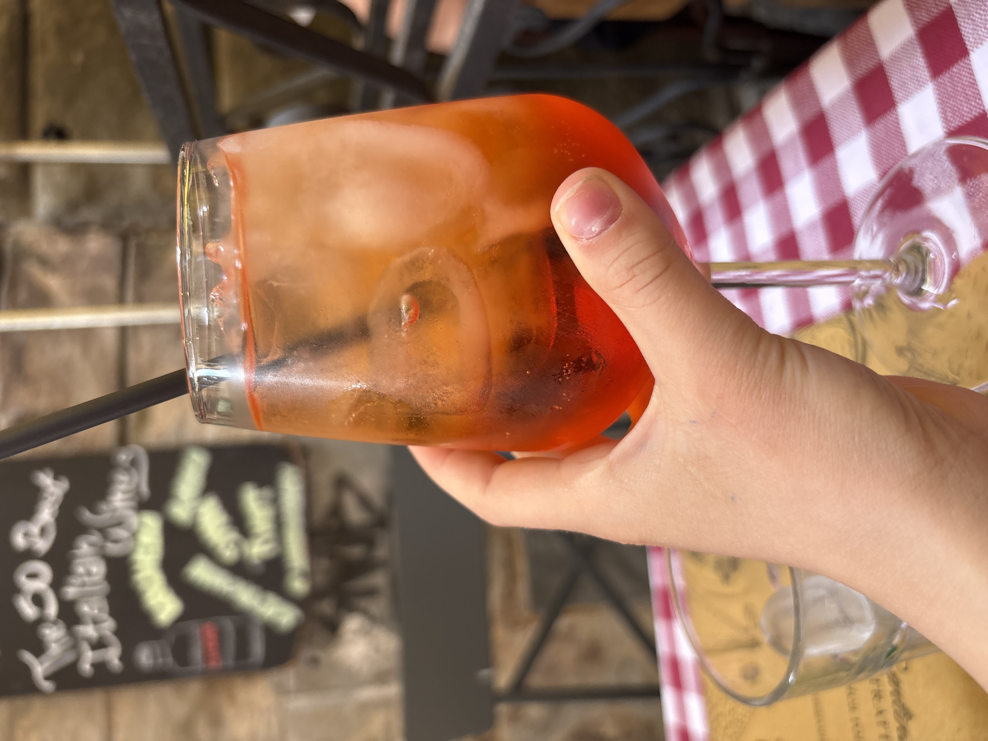 A first-person, slightly rotated shot shows a person's right hand holding a vibrant orange-colored drink in a clear, wide glass. The drink contains ice cubes and a black straw, resembling a refreshing cocktail such as an Aperol Spritz. The person's thumb and forefinger are visible, with neatly trimmed fingernails.
The setting appears to be an outdoor cafe or restaurant, characteristic of Rome, Italy. A classic red and white checkered tablecloth covers a table to the right, where another empty or near-empty wine glass with ice is placed.
The background is softly blurred but reveals elements typical of an urban outdoor dining area: a light-colored, possibly stone or tiled ground, and dark metallic furniture frames. On the far left, a dark sign, likely a chalkboard, displays blurry white and yellow chalk writings. Distinguishable details on the sign include what appears to be a price, "8,00", positioned vertically, and other indistinct words and shapes, possibly menu items or decor. The bright, natural lighting suggests it is daytime, perhaps late afternoon, a popular time for enjoying such drinks.