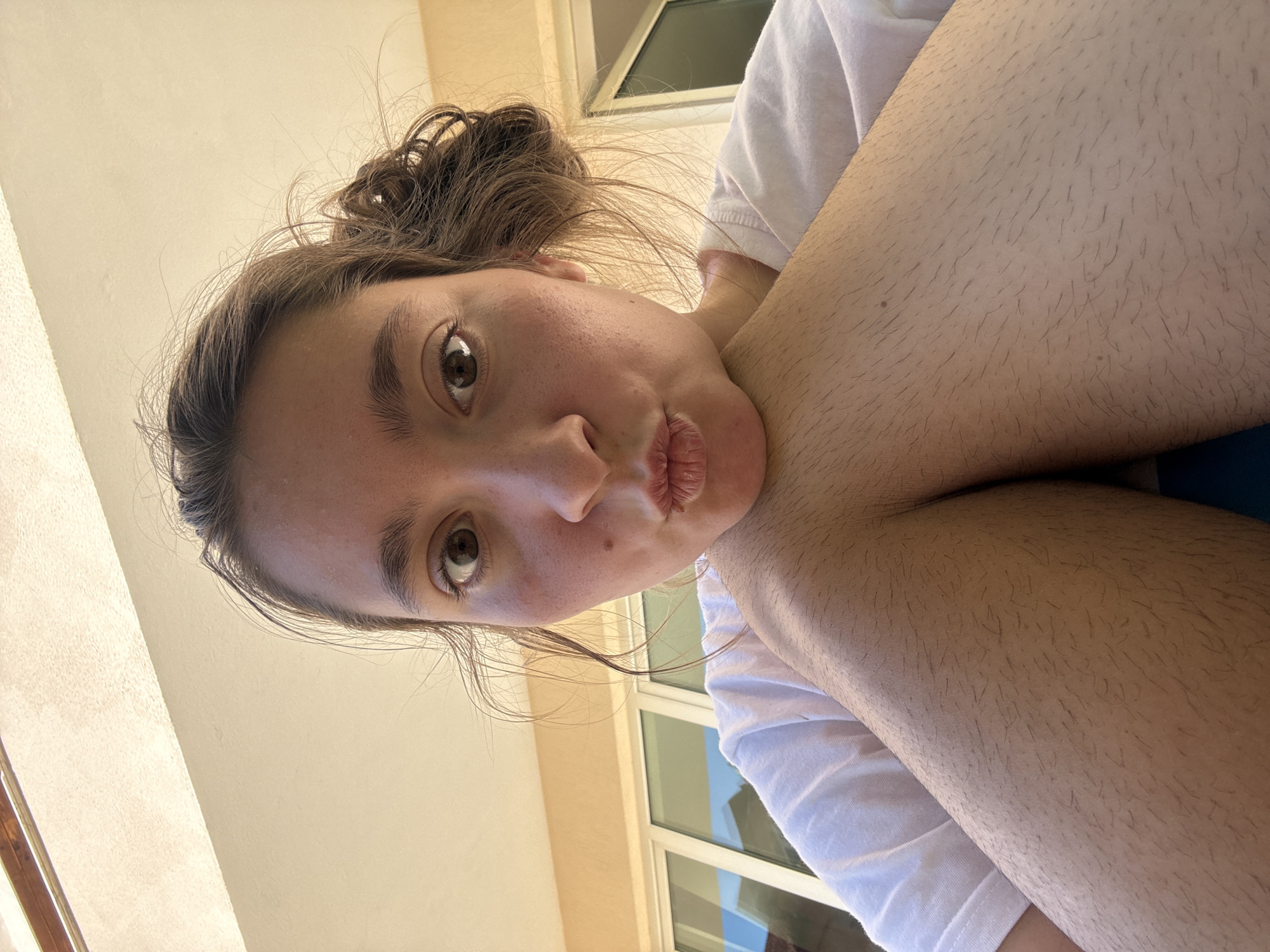 An extreme close-up, low-angle selfie features a young woman with light brown hair pulled up in a messy bun and dark brown eyes. She is wearing a white t-shirt and her arms are bent, with her left elbow resting on her right knee, creating a V-shape. Her lips are pursed, as if for a kiss. The background shows a building with pale yellow walls and large windows, suggesting an outdoor location with natural light. The scene is captured from the perspective of someone lying down, looking upwards at the woman.