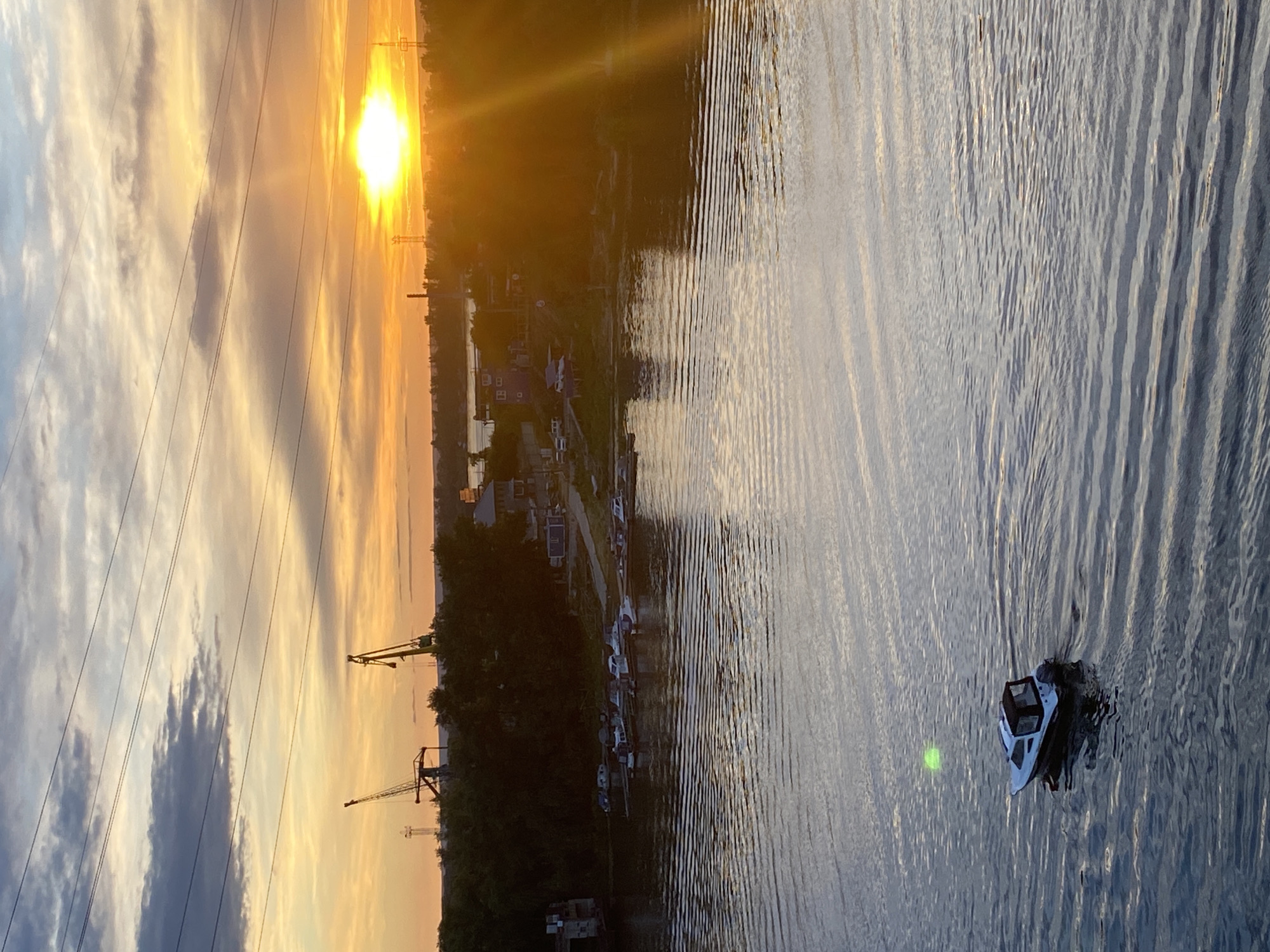 This is a landscape photograph taken outdoors, likely during the evening as the sun is setting. The scene is dominated by a body of water, with ripples creating a textured surface that reflects the warm hues of the sunset and the sky. A small boat with a cabin is visible on the water, moving away from the viewer.
Along the shoreline, there is a dense line of trees and some buildings, appearing to be part of a settlement or industrial area. Power lines stretch across the sky, intersecting with the sunset's light. In the background, industrial cranes are also visible, suggesting a port or industrial zone. The sky displays a dramatic gradient of colors, from bright yellow and orange near the sun to softer blues and grays with scattered clouds.
The overall mood is serene and tranquil, characteristic of a sunset over water. The time of day is clearly evening, and the weather appears clear with some cloud formations. The location cues suggest an industrial waterfront area, possibly in Novokuybyshevsk, Russia, given the context provided. No text is visible in the image.