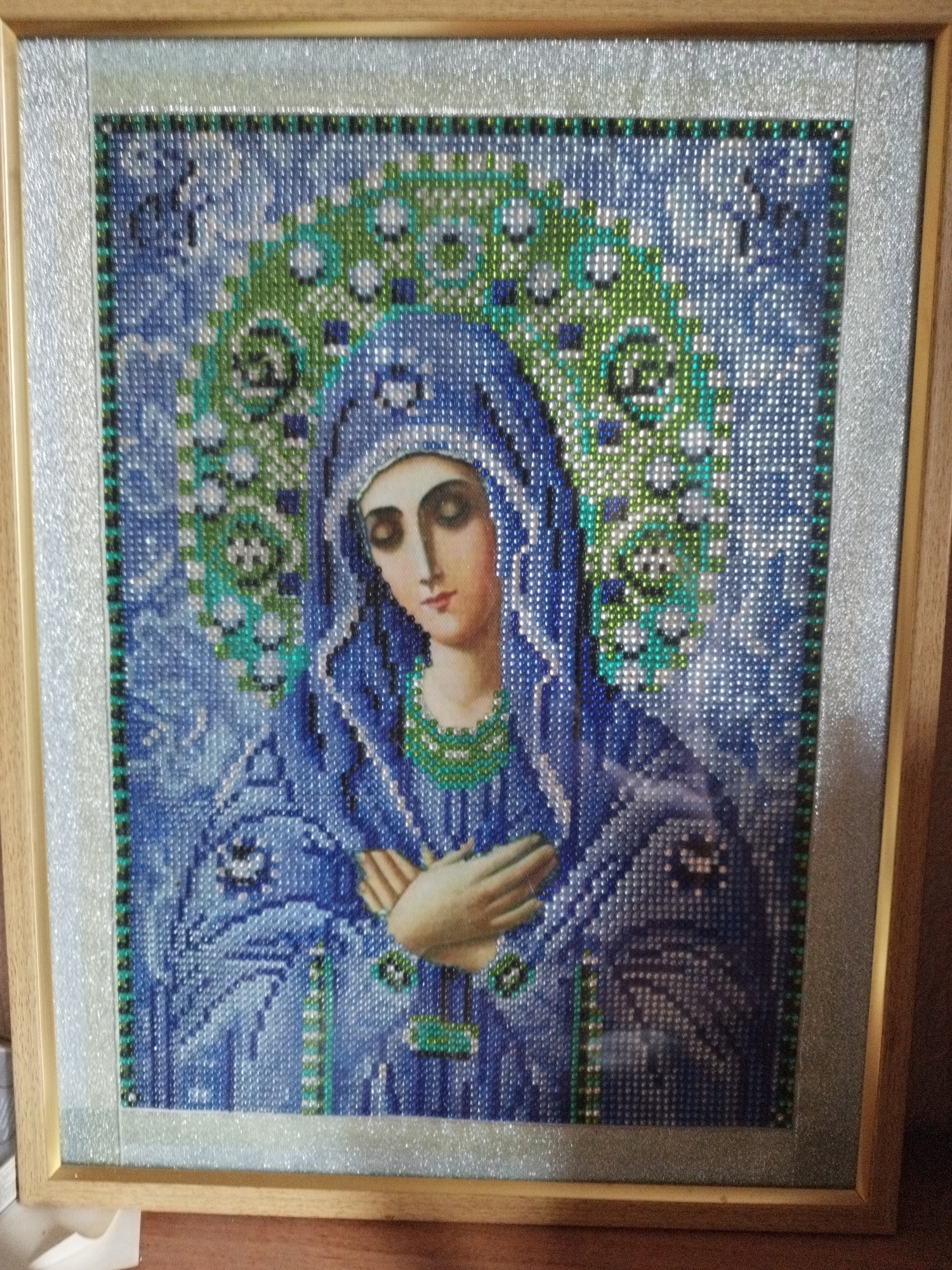 This is a framed picture of a religious icon, specifically a depiction of the Virgin Mary. The image is created using a mosaic of small beads, giving it a textured, pixelated appearance. The Virgin Mary is rendered in shades of blue, with her head covered by a blue veil. She is looking downwards with a serene and somber expression, her hands crossed over her chest. Behind her is a halo-like circular design in shades of green and blue, reminiscent of a peacock feather motif. The background is a light blue, with some darker blue and white accents that suggest fabric or drapery. The beadwork is meticulously arranged, forming distinct lines and shapes that define the figures and decorative elements. The artwork is housed in a simple wooden frame with a silver, glittery mat surrounding the beaded image. The lighting in the photograph is warm, suggesting an indoor setting, possibly a home or a place of worship. There is no visible text in the image.