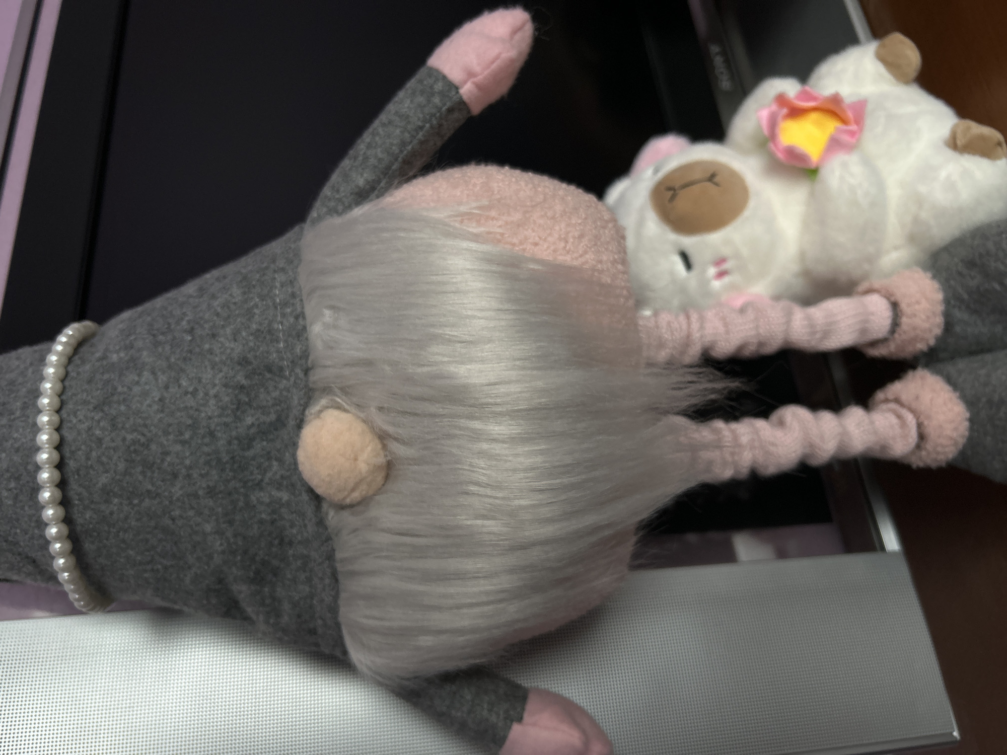 A close-up image shows two plush toys resting on a surface with a metallic finish. The primary subject is a gray gnome-like figure with a long, flowing white beard, a pink cap, a visible nose, and a string of pearls around its "neck". Beside the gnome is a smaller, white plush sheep with a pink flower attached to its head. The background includes a dark, reflective surface that appears to be part of an electronic device, possibly a monitor. The description is forensic and matter-of-fact, containing only the requested information. There is no mention of the city of Saint Petersburg, Russia, as there's nothing in the image to suggest that location.