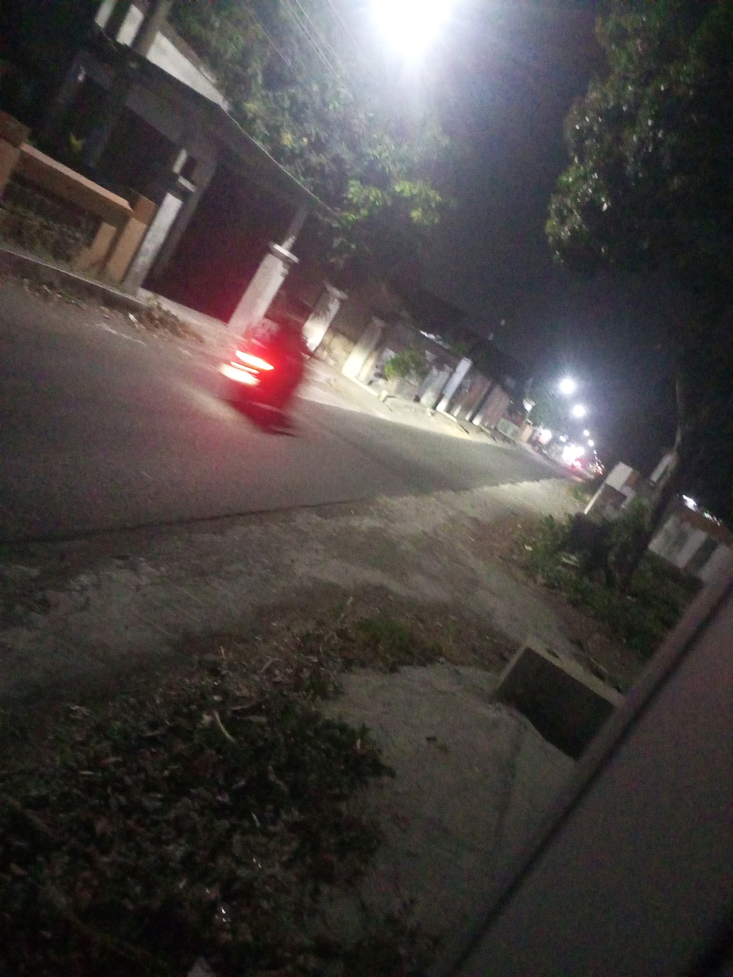 The image captures a nighttime street scene in Ngasem, Indonesia, characterized by low lighting and motion blur. A motorcycle with illuminated red taillights is moving from left to right across the asphalt road, its motion creating streaks of light. The road appears wet, possibly from recent rain, reflecting the streetlights. On the left side of the road, a sidewalk is visible, lined with debris, leaves, and sparse vegetation. Adjacent to the sidewalk are structures, likely residences or shops, with darkened facades and some illuminated windows. Further down the street, more streetlights are visible, creating pools of light on the road and illuminating the surroundings. The overall environment suggests a residential or commercial area in Indonesia at night.