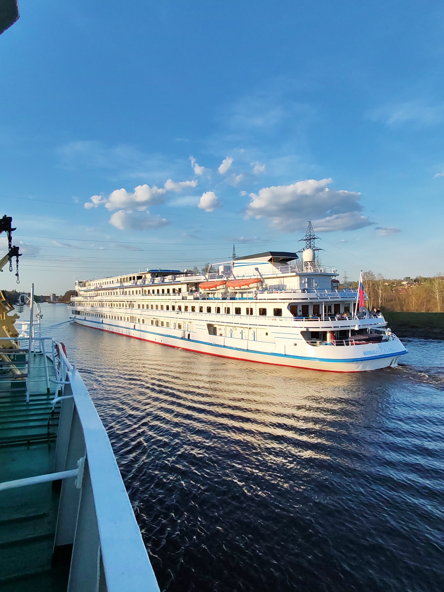 A daytime, eye-level shot captures two vessels traversing a waterway in Базарово, Russia. The larger vessel, painted white with red and blue accents, is the focal point. It features multiple decks with numerous windows and what appears to be a Russian flag waving near the stern. On the bow of the ship reads "LEONID KRASIN." The water surface reflects the light and the colors of the vessels and the sky. The foreground shows the side of a smaller vessel, its colors predominantly green and white. Some cranes are visible on the side of the smaller ship. A portion of the sky is visible, dotted with white clouds. The shoreline of Базарово is visible in the background, featuring trees and buildings. Overhead, power lines stretch across the sky. The boats appear to be navigating adjacent to each other.