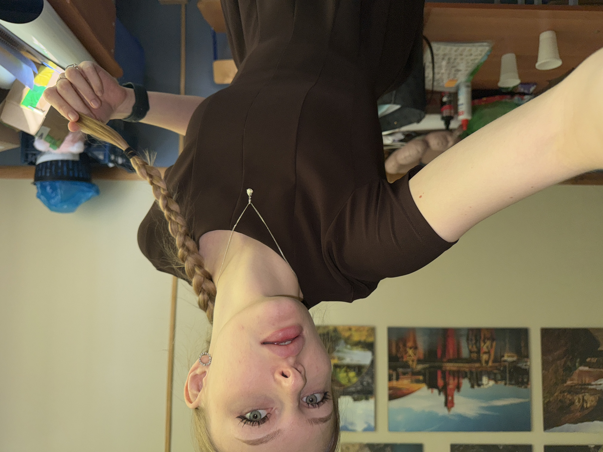 A woman is facing the camera in an upside-down orientation. She has blonde hair in a braid, fair skin, and light eyes. She is wearing a brown dress and a delicate necklace. Her left arm is extended upwards, holding the end of her braid. Her right arm is extended to the right. She is wearing a dark wristband. Behind her are several small landscape images, including what appears to be an upside-down view of the Perm State Art Gallery in Perm, Russia. Other objects visible include a desk with various items.