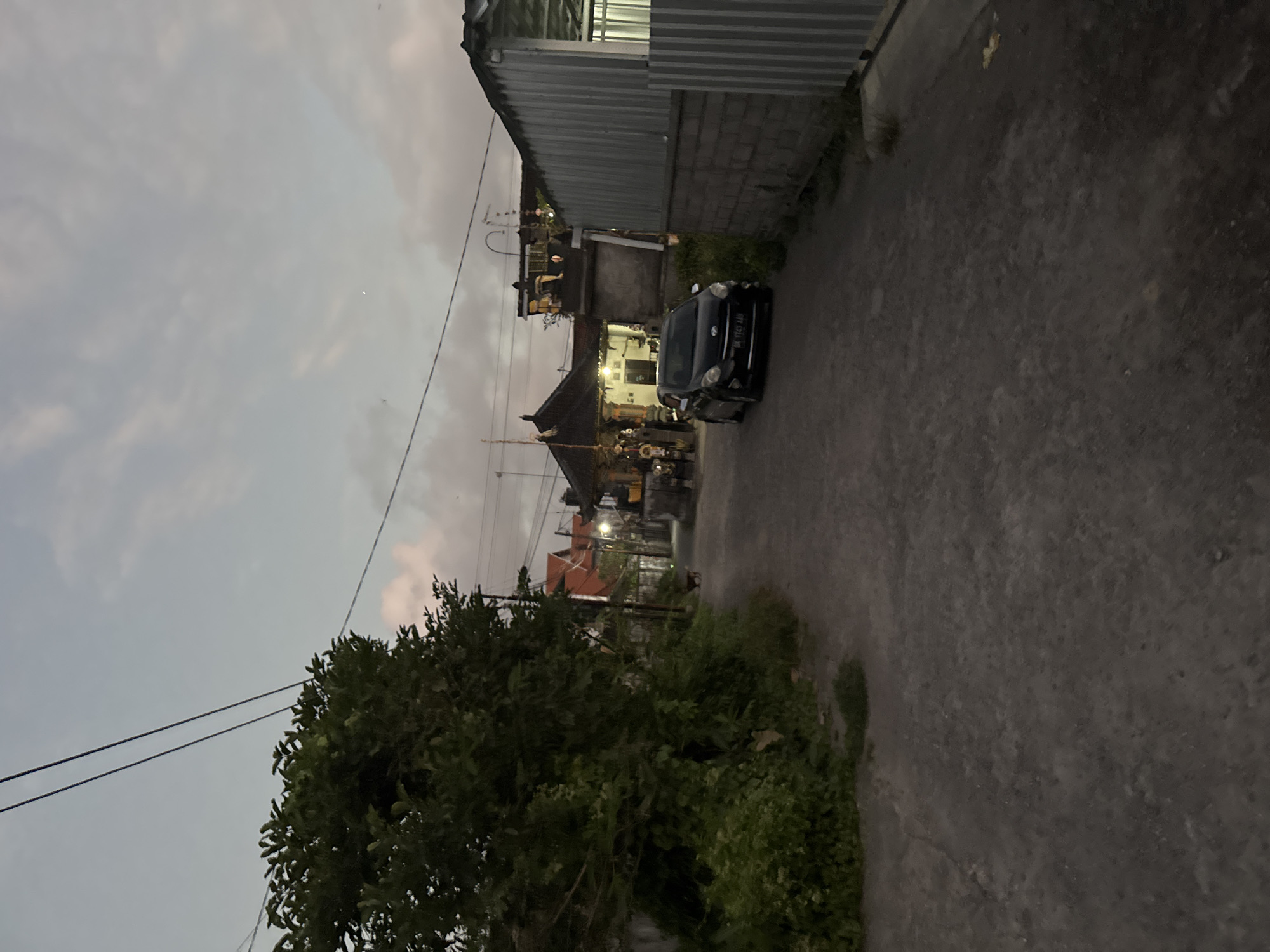 The image depicts a narrow, unpaved road in a residential area, likely in Sukawati, Indonesia, during the twilight hours. Overhead power lines traverse the sky. On the left, a dense cluster of lush green foliage dominates the foreground. Further down the road, a black car is parked, facing away from the viewer. On the right side of the road, a building constructed with grey brick and corrugated metal roofing is visible. The background features traditional Balinese-style houses with dark, sloped roofs and decorative elements, some illuminated by interior lights. The overall atmosphere is calm and subdued, characteristic of the early evening in a rural or semi-urban Indonesian setting.