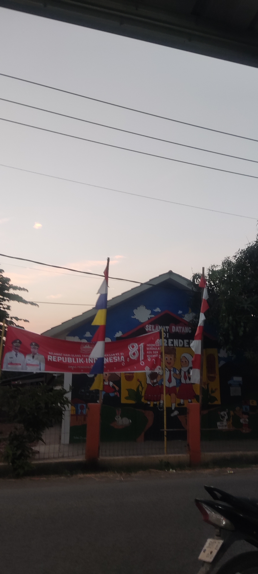 A banner celebrating Indonesia's 81st Independence Day is displayed in front of a building, possibly a school, in Getrakmoyan, Indonesia. The banner features images of two men in uniform, the text "SELAMAT HARI ULANG TAHUN KE 81 REPUBLIK INDONESIA" and "INDONESIA BERDAULAT ADIL DAN MAKMUR". The building's facade is decorated with colorful murals of children. Two flags, one with red and white stripes, and another with red, white, blue, and yellow segments, fly near the entrance. A motorcycle is parked on the street in the foreground. The sky is overcast and the lighting suggests it is either early morning or late afternoon.