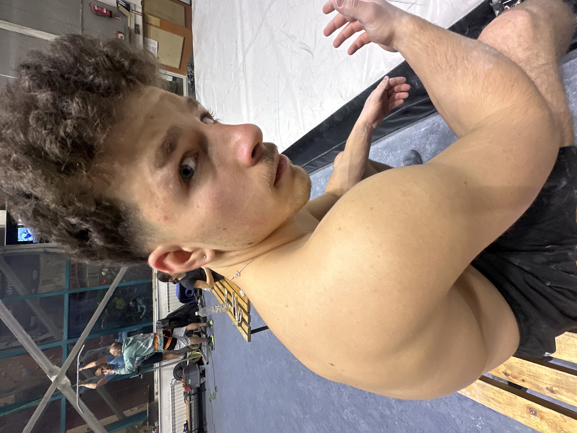 A young, shirtless man with curly brown hair is sitting on a wooden pallet in what appears to be a climbing gym. He is looking at the camera. There is a climbing wall in the background, and other people can be seen in the gym. It is not possible to determine the exact location within Sofia, Bulgaria from the image alone.