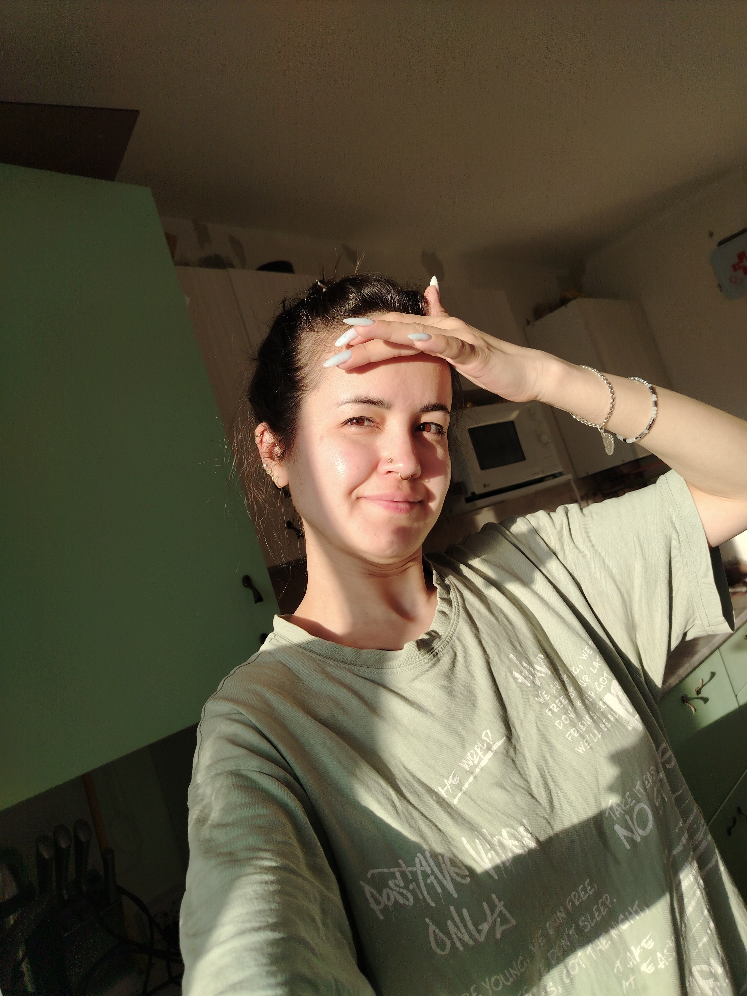 The media file presents a well-lit selfie of a young woman in what appears to be a kitchen in Noviy, Russia. Strong, warm sunlight streams into the room, suggesting either morning or late afternoon, creating bright highlights on her face and a distinct shadow from her right hand as she shields her eyes.
The woman has dark hair styled up, revealing her forehead. She has fair skin, multiple ear piercings, and a nose stud. Her long fingernails are painted white or a very light color. She is wearing a light sage green t-shirt adorned with various white graphic texts. On her left wrist, she wears a silver chain bracelet and another beaded bracelet.
The text visible on her shirt includes phrases like "THE WORLD," "Pastime Always," "ONLY," "BE YOUNG, WE RUN FREE," "WE DON'T SLEEP," and "GOT THE NIGHT." On the right side of her shirt, phrases such as "WE ARE, WE'D," "FREEST UP LATE," "FRIENDS," "DON'T STOP, GOT," "WE'LL BE PLAYING," "TAKE IT EASY," and "NO" are also discernible.
The background shows green kitchen cabinets or walls on the left, while the right side features white cabinetry and a microwave oven, confirming the indoor kitchen setting. The woman exhibits a relaxed, slightly smiling expression, conveying a sense of contentment or casual ease despite the bright sunlight.