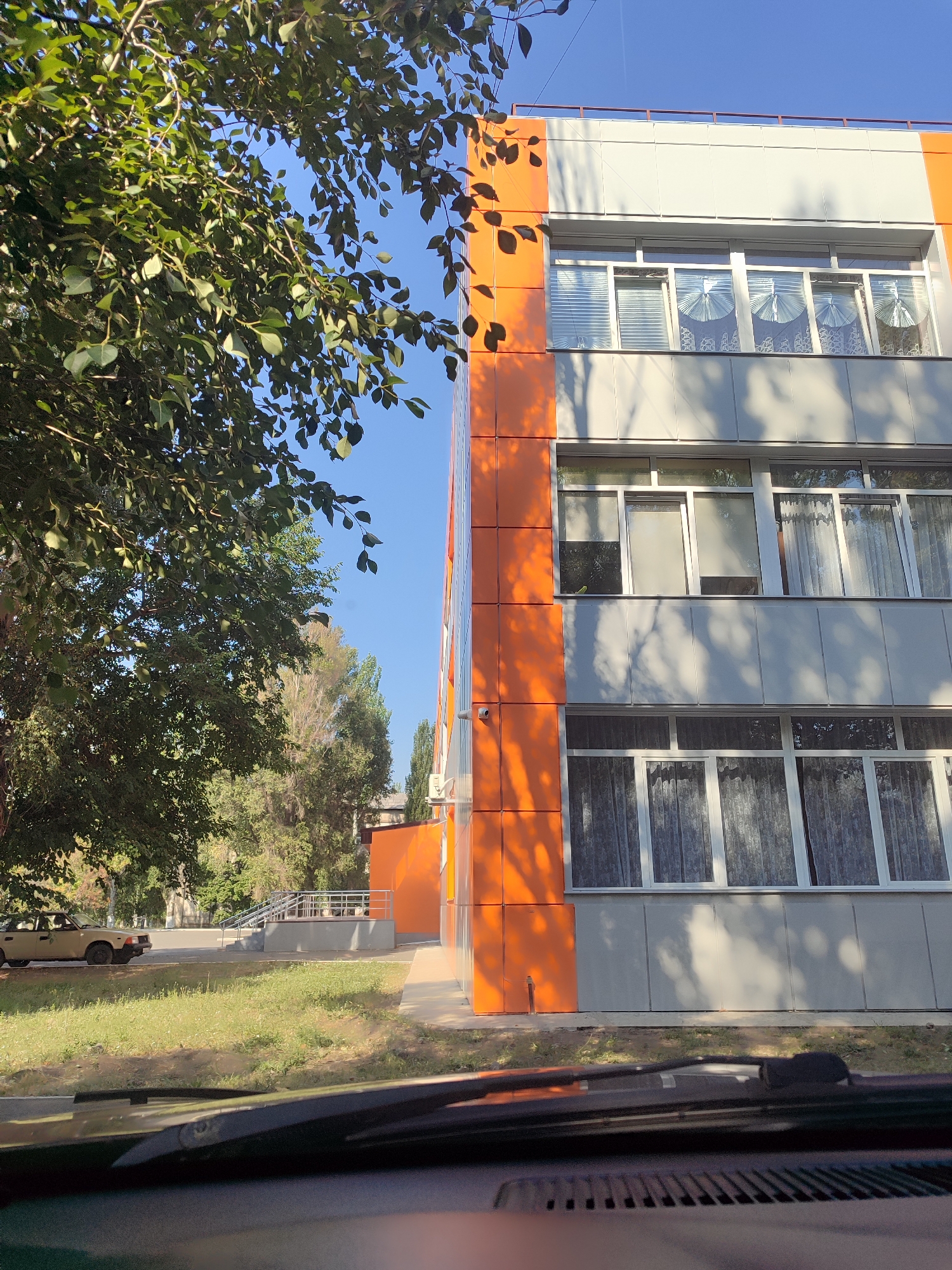 The image is a photograph of the side of a modern building with orange and grey paneling, featuring multiple rows of windows. The building is located in a city, with trees and vegetation visible to the left. A light-colored sedan is parked on the left side of the frame. In the foreground, the dashboard and windshield of a vehicle are visible, suggesting the photo was taken from inside a car. The sky is clear and blue.
