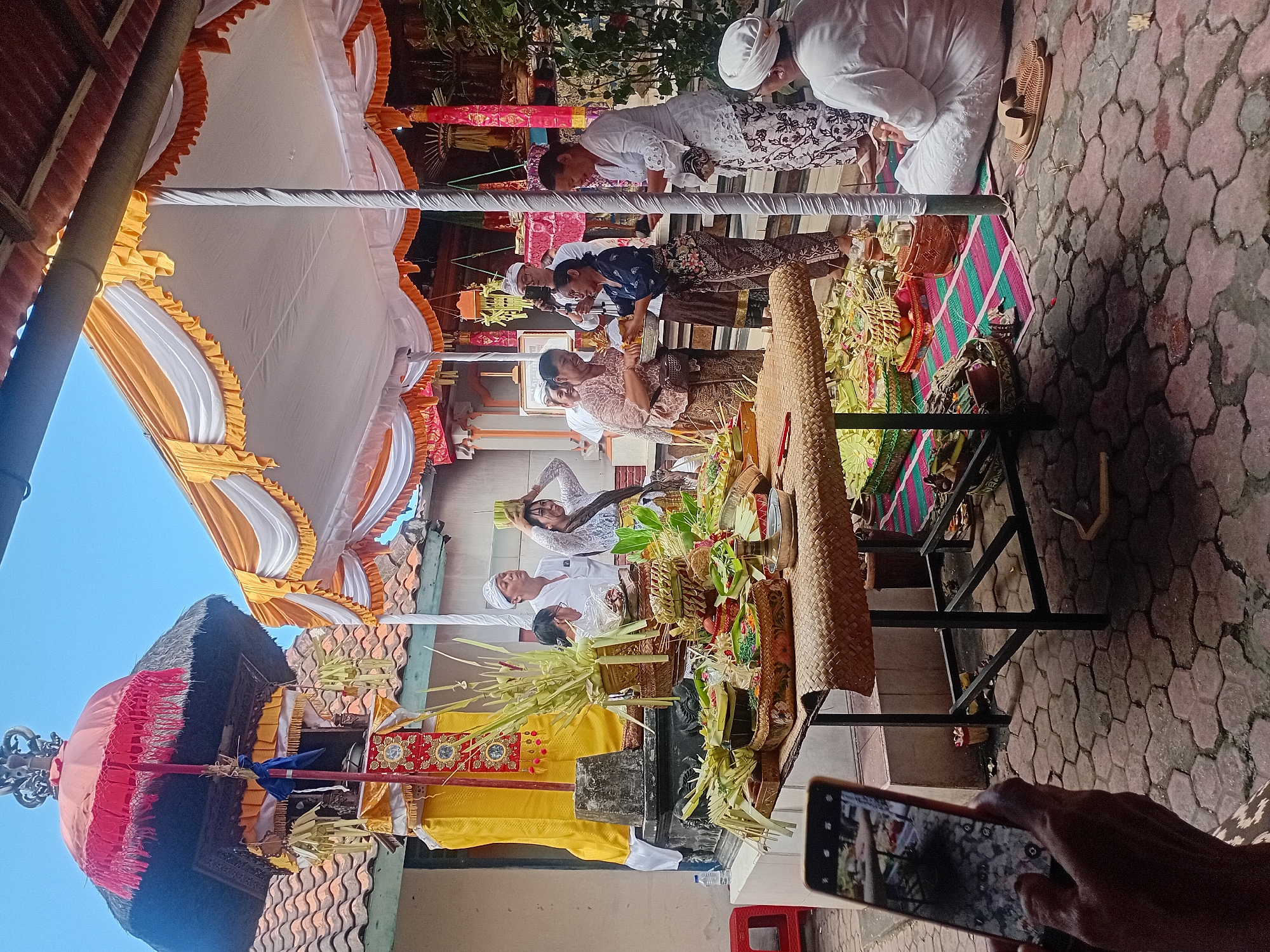 The image depicts an outdoor religious ceremony taking place on a paved courtyard, likely in Kuta, Indonesia. The scene is set under a temporary canopy adorned with white and orange fabric. In the background, a traditional Balinese temple structure with a thatched roof and an ornate red umbrella is visible. A table laden with offerings, including woven baskets filled with food, fruits, and decorative items made of palm leaves, is prominently displayed in the foreground. Several individuals, dressed in traditional attire, are participating in the ceremony. Some are holding offerings, while others appear to be observing or assisting. One person in the foreground is capturing the event with a mobile phone. The atmosphere suggests a communal and spiritual gathering.