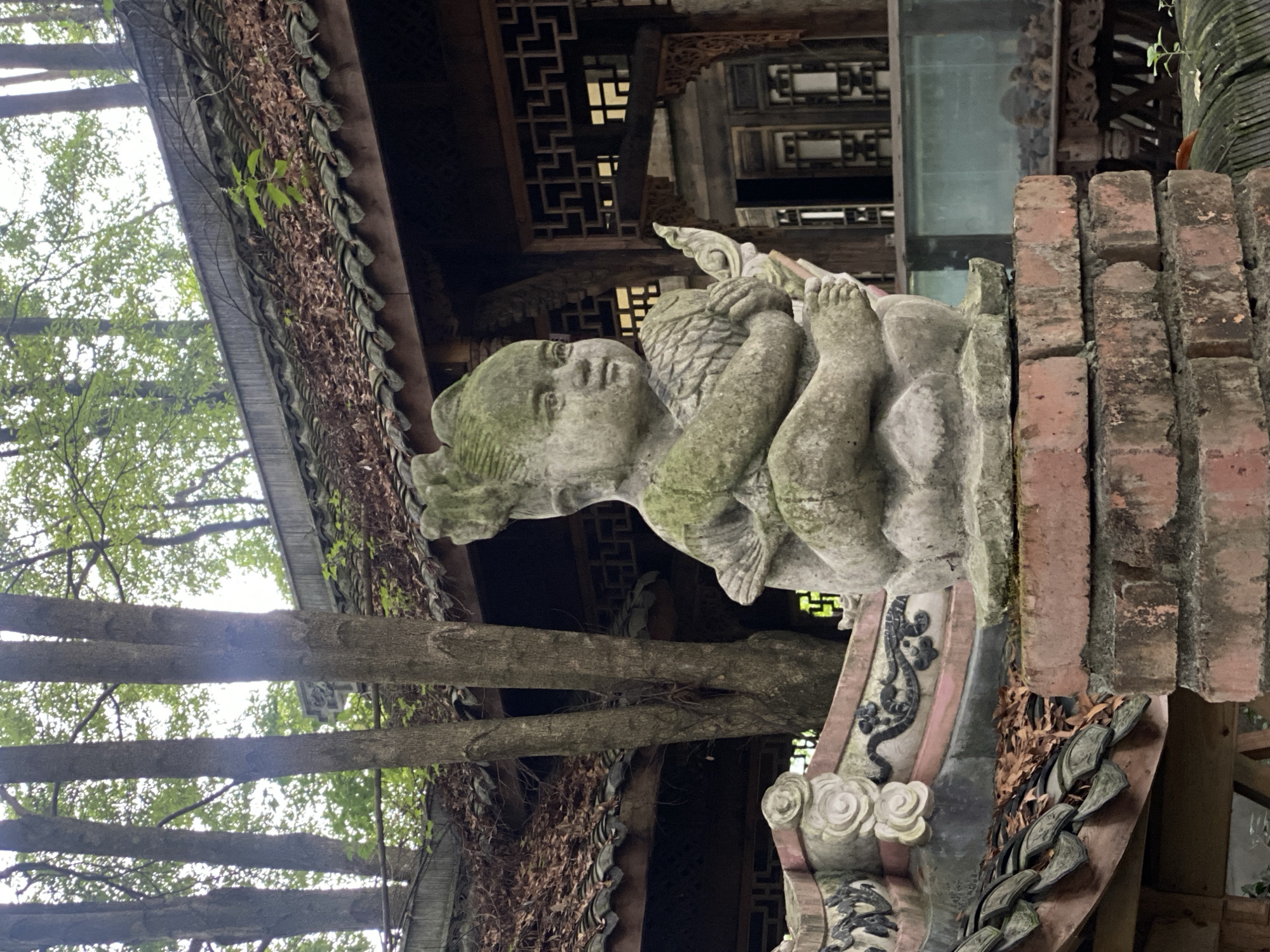 The image depicts a stone sculpture of a child, possibly an infant, in a seated or reclining pose, with its legs and arms visible. The sculpture is weathered and covered in moss, suggesting it is outdoors and has been exposed to the elements for some time. The child appears to be holding an object, possibly a lotus flower or a similar symbolic item, with its hands.
The sculpture is positioned on a brick structure, which is part of a larger, traditional Chinese architectural setting. In the background, there are wooden beams and intricate latticework, indicative of a temple, ancestral hall, or historical building. The roof tiles are visible, some of which are covered in dried leaves and vines, further emphasizing the outdoor environment and the passage of time. Trees with lush green foliage are visible to the left of the sculpture, and branches extend across the frame, suggesting a natural, possibly overgrown, setting.
The lighting appears to be natural daylight, casting soft shadows. The overall mood is one of serenity and history, with the weathered sculpture and traditional architecture evoking a sense of age and cultural heritage. There is no visible text in the image. The location is identified as Chongzhou City, China. The scene appears to be a quiet, possibly sacred, outdoor space.