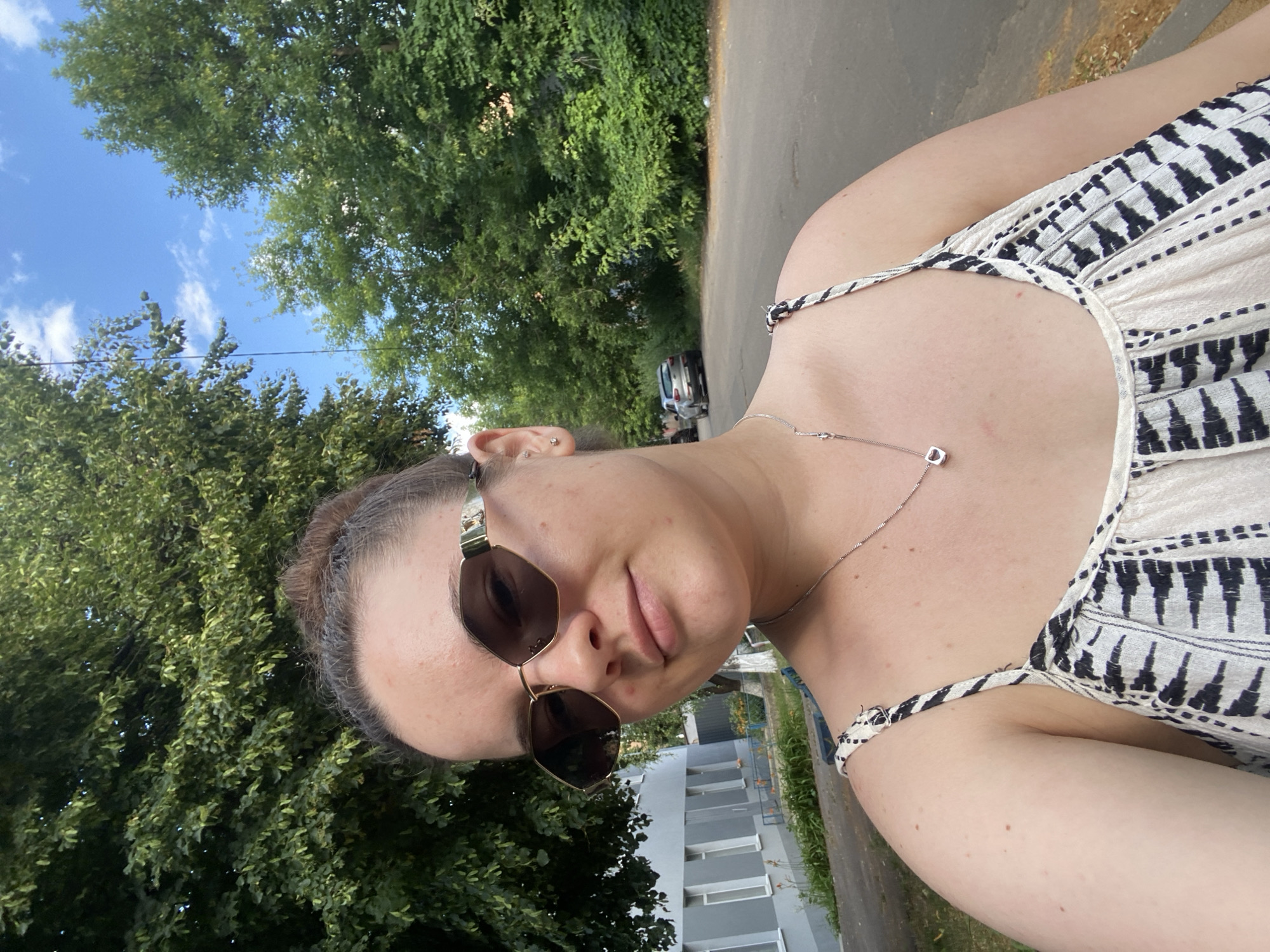 The media file is a selfie taken outdoors on a sunny day in Poltava, Ukraine. The subject is a person wearing sunglasses and a white and black patterned dress with spaghetti straps. They are also wearing a silver necklace with a small, square pendant. The background features lush green trees, a paved road, and a modern building with multiple stories and balconies. The sky is blue with scattered white clouds. The overall mood appears relaxed and casual. The time of day seems to be daytime, given the bright sunlight.