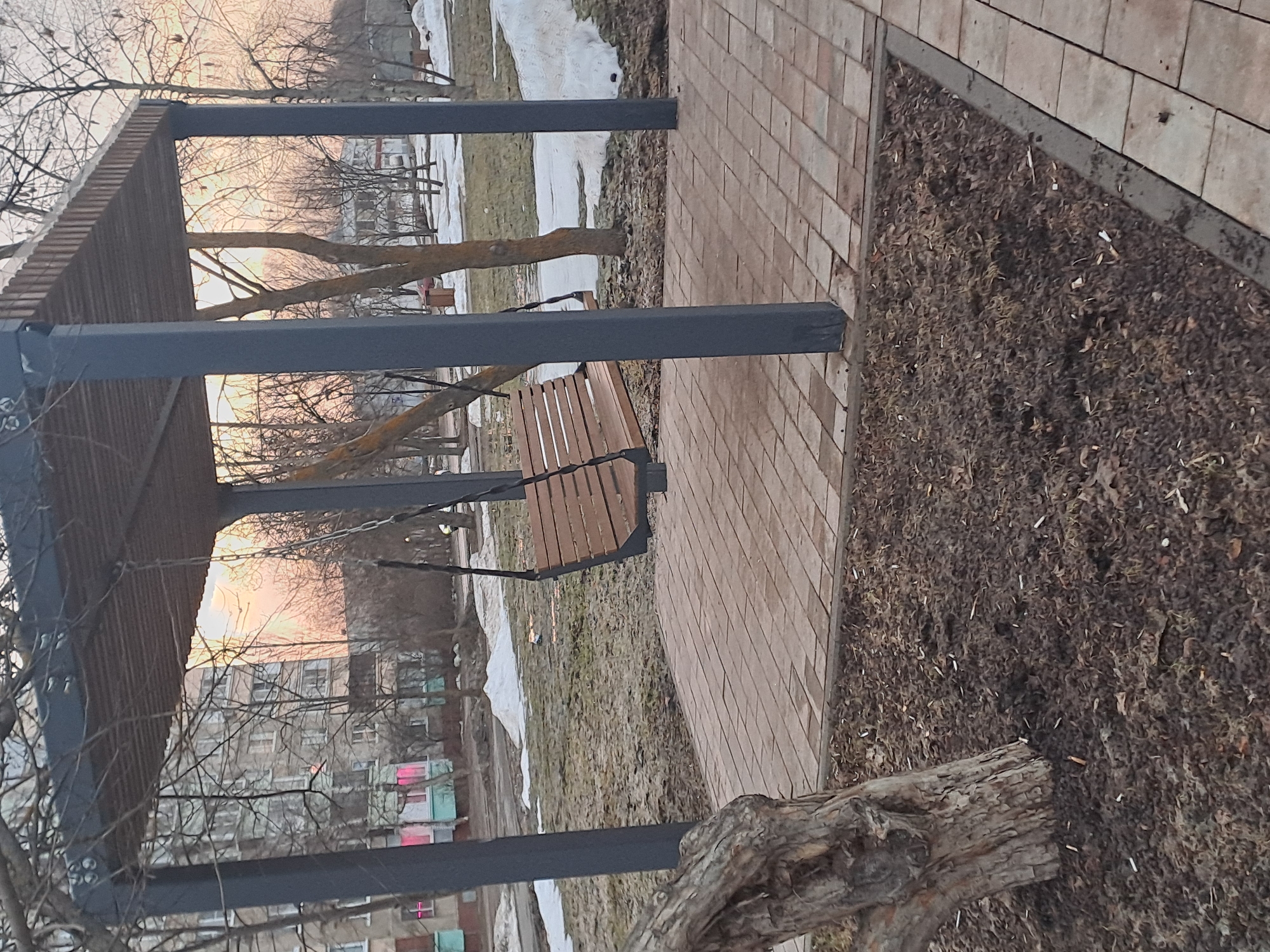 An outdoor scene in Elektrostal, Russia, depicts a public amenity during what appears to be late winter or early spring, characterized by bare deciduous trees and patches of melting snow. The central feature is an unoccupied, modern wooden swing bench, constructed from horizontal slats, suspended by metal chains from a dark grey, rectangular-posted pergola with a slatted roof structure. This pergola stands on a platform of light-colored rectangular paving stones.
In the immediate foreground, to the bottom left, a gnarled tree trunk with rough, light brown bark is visible. The ground surrounding the paved area is primarily dark brown earth, covered with fallen leaves and scattered detritus, including small white fragments resembling cigarette butts.
Beyond the paved area, sparse, dry grass and irregular patches of melting snow cover the ground. Numerous bare trees, some with lighter trunks, are dispersed throughout the midground. A dark brown trash bin is partially visible among these trees on the left side.
In the background, a multi-story, light-colored apartment building stands on the left. Several of its windows display a distinct bright pink or magenta glow from within, and the building's lower section features light blue-green details. To the right, more bare trees obscure the distant view, with a very blurry, light-colored figure partially visible near a tree. The sky, seen through the tree branches, exhibits a pale orange and pinkish hue, consistent with either sunrise or sunset. No identifiable individuals are actively engaged in the immediate scene.