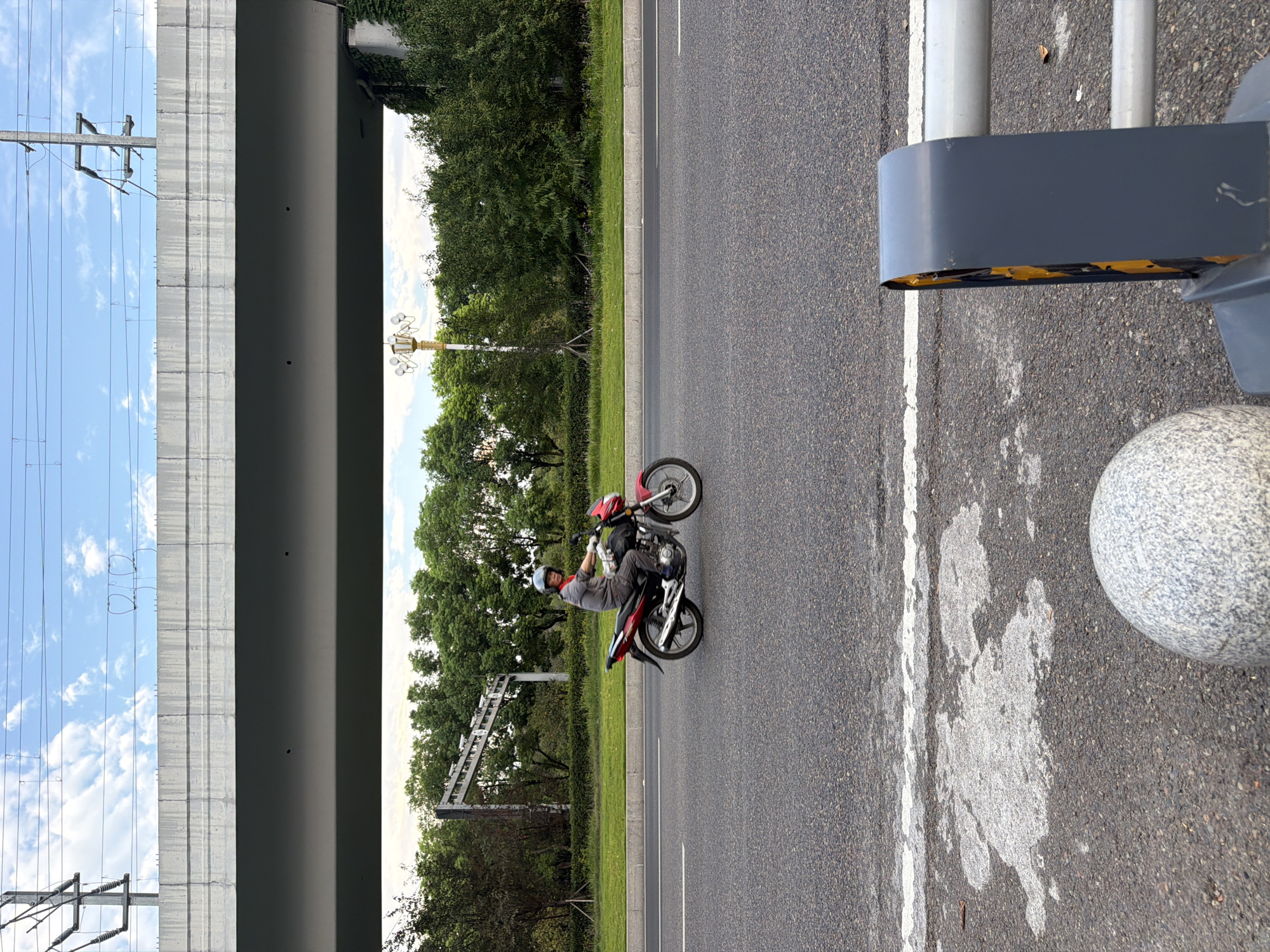 A person is riding a red motorcycle on a paved road in Jiangjin, China. The rider is wearing a gray long-sleeved shirt, gray pants, a helmet, and gloves. The motorcycle is a mid-size model. The road is divided by white lines, and there is a grassy median to the left of the road. In the background, there are trees and an elevated overpass with overhead power lines. The sky is partly cloudy. In the foreground, there is a gray sphere and part of a dark gray protective barrier.