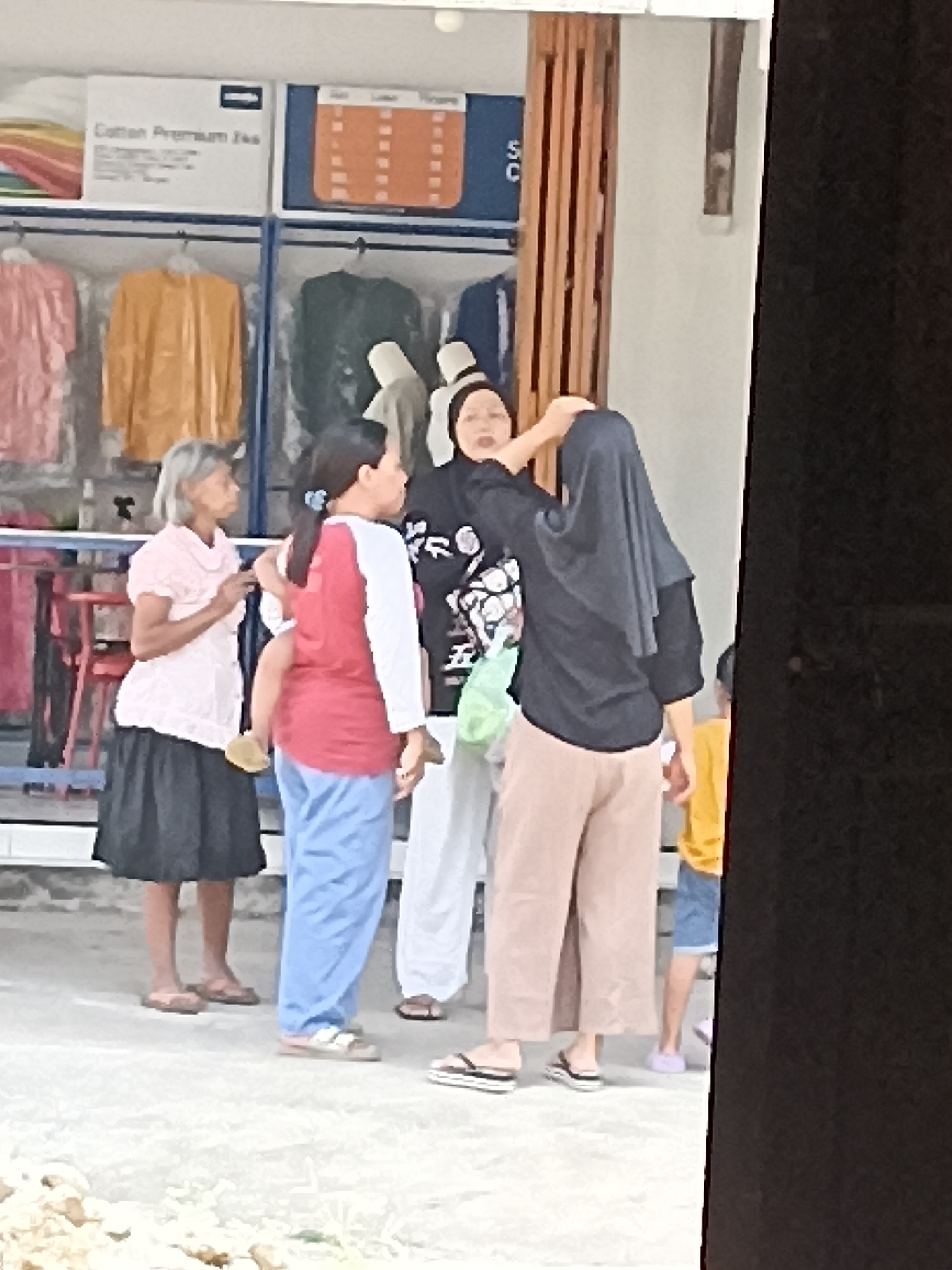 In the city of Pulisen, Indonesia, a group of individuals is gathered near a storefront. On the left, an elderly woman with grey hair, wearing a light pink short-sleeved shirt and a black skirt, stands looking to the left, holding a yellow slipper in her right hand. Next to her, a woman wearing a red and white long-sleeved shirt and light blue pants stands facing away from the camera, with her right arm around a child who is partially visible. Another woman, wearing a black hijab and a black long-sleeved shirt with a white pattern on the front, is being assisted by someone with their hijab. She is wearing light brown pants. To the right, a child in a yellow t-shirt and blue shorts stands. Behind them, clothing items are displayed on racks. The ground is a light-colored, textured surface. The scene appears to be outdoors, possibly in a market or commercial area, with a black rectangular object partially obscuring the right side of the image.