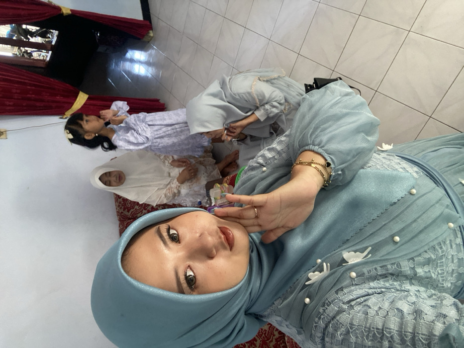 A domestic scene in Tambakrejo, Indonesia, features four individuals. In the foreground, a young adult woman in a light blue hijab and a matching light blue dress with sheer puffed sleeves, pearl, and butterfly embellishments poses for a selfie. She wears a gold ring on her right ring finger and a gold bracelet with black heart charms on her right wrist, which is raised with the palm open and fingers slightly splayed, touching her jawline. Her expression is a subtle smile directed at the camera. Behind her, seated on a reddish-brown patterned couch, an older woman wears a white hijab and a white patterned dress, looking generally forward. To her right, a young girl stands, wearing a light purple patterned dress and a white bow in her dark hair, with her hands clasped at her chest and looking towards the right. Further right, an adult woman sits on the tiled floor, dressed in a light blue/grey hijab and matching garment, leaning forward with her right hand near her mouth. The room has plain white walls and a light-colored square-tiled floor. Red curtains with gold ties frame an opening on the right, revealing a glimpse of an exterior or another room with a motorcycle visible through the doorway. Bright light enters from the right side, casting shadows on the floor.