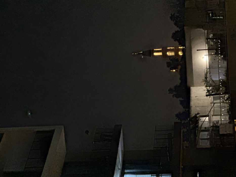 An upside-down night view captures city buildings and a illuminated church tower. The tower features a pointed spire and visible clock face. The sky is overcast and dark. Buildings with windows and balconies are on the right side of the image. Silhouetted structures appear in the lower portion of the composition.