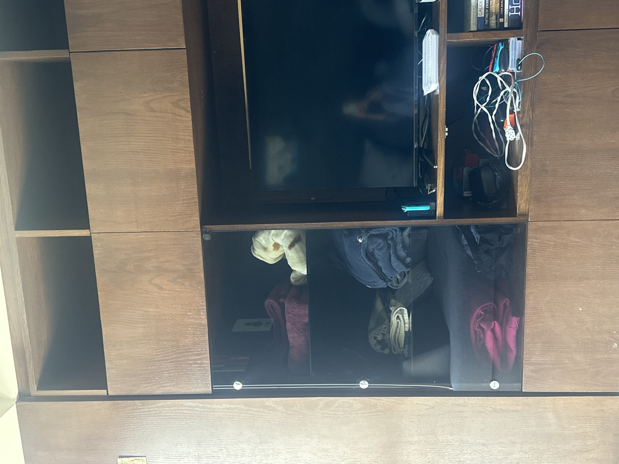 The image depicts an interior scene, specifically a multi-tiered wooden shelving unit or wall cabinet located in a residence in Huixquilucan de Degollado, Mexico. The unit is constructed from medium-brown wood with a visible grain.
The upper section features two empty, open cubby shelves. Below these, the unit is divided into several compartments. The left section consists of a glass-fronted cabinet with three internal shelves. The top shelf holds a stack of folded burgundy towels and a light-colored, possibly cream or yellow, folded textile. The middle shelf contains a stack of light-colored, patterned folded textiles and a dark blue folded blanket. The bottom shelf displays a dark pink folded textile, possibly a blanket or clothing. The glass door of this cabinet is secured with three visible circular fasteners along the left edge.
The right side of the unit features an electronic entertainment setup. A black television screen, displaying a faint reflection of a person holding a mobile device, occupies a central position. The television is identified as "JVC." Below the TV, a wooden shelf holds a light blue Nintendo Switch console standing vertically on the left, and a white rectangular container on the right. Below this shelf, an open compartment contains a dense cluster of entangled white, black, and multicolored cables, various electronic adapters, and a red gaming controller. To the right of the cable compartment, a stack of books is visible, with titles such as "GAUDI," "THE END OF EVERYTHI," "CASA BATLLO," and "barcelona." The lower portion of the unit comprises closed wooden cabinet doors. The top edge of a light-colored ceiling is visible at the very top of the frame. No individuals are present in the scene, save for the reflection on the television screen.