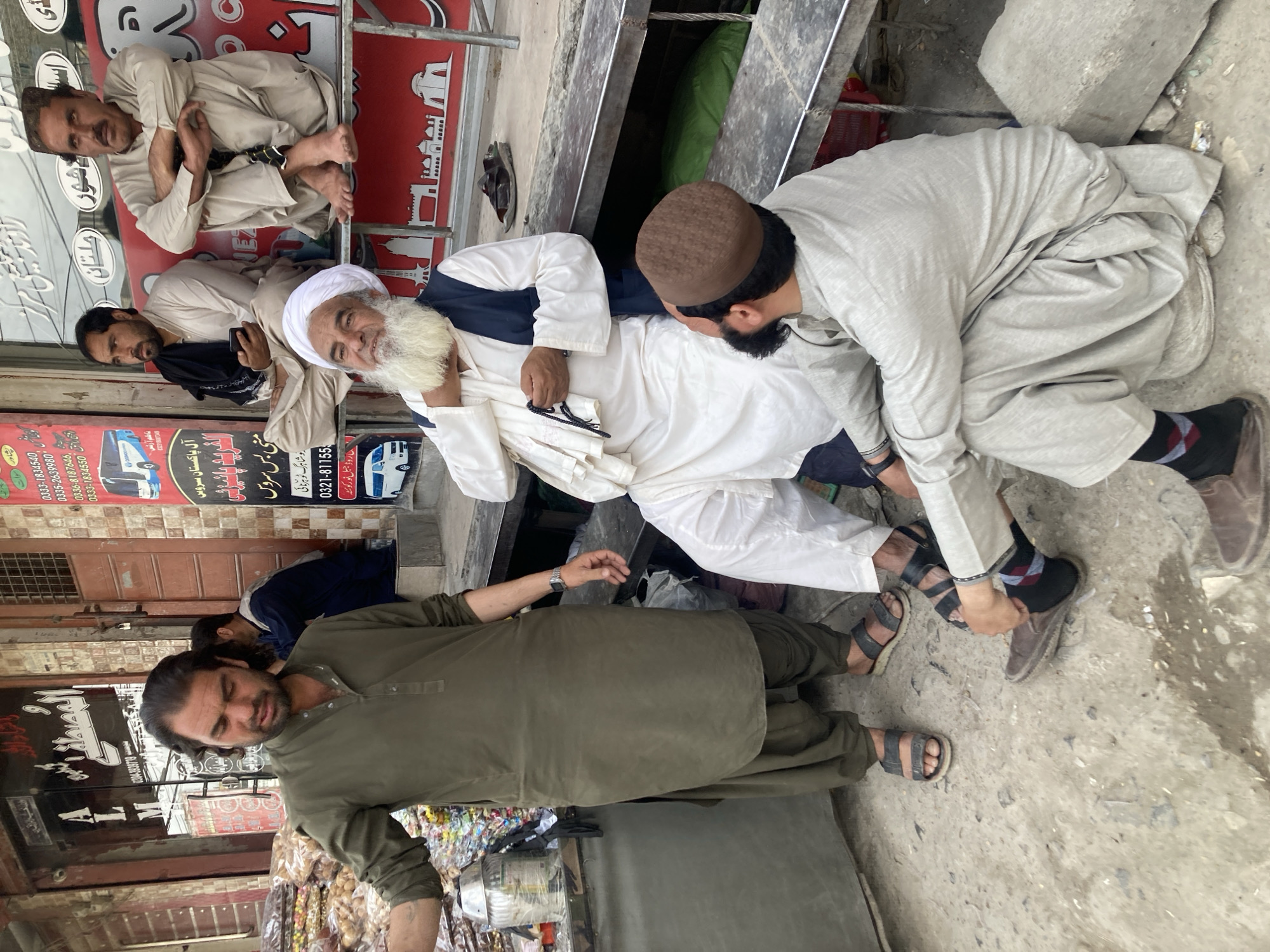 A street scene in Quetta City Tehsil, Pakistan, captures a group of men gathered in what appears to be a busy commercial area during daytime. The lighting suggests an overcast sky or diffused sunlight.
In the foreground, an older man with a prominent white beard and white turban, wearing a white shalwar kameez and a dark vest, sits on a raised platform or bench. He is smiling and holds a string of prayer beads in his hand. A younger man, wearing a light-colored shalwar kameez and a brown prayer cap, bends over him, gently holding his foot or ankle, suggesting an act of care or assistance. This younger man wears patterned socks and brown shoes.
To the left of the central interaction, another man in a light brown/beige shalwar kameez sits on a higher ledge, cross-legged, looking directly at the camera with a relaxed expression. Below him, another man in similar attire sits on the ground, his back slightly turned, looking forward. Further to the left and slightly in front, a man in an olive green shalwar kameez stands, wearing sandals and a watch, his gaze directed away from the central group. A partially visible figure stands behind him.
The ground is concrete and appears somewhat worn. The background features various shopfronts with visible Urdu text on signs and banners. Notable texts include "All Pakistan Service," "Mini Bus Service," and specific routes like "Nowshahrag to Harnai," and a location "Maa Road Jabal Noor Quetta," confirming the city. Other shop names like "Al-Mustafa" and "Aai Shawar (Multan)" are also visible, along with multiple phone numbers. The scene depicts a casual street gathering, possibly related to travel services given the signs, with one man tending to another.