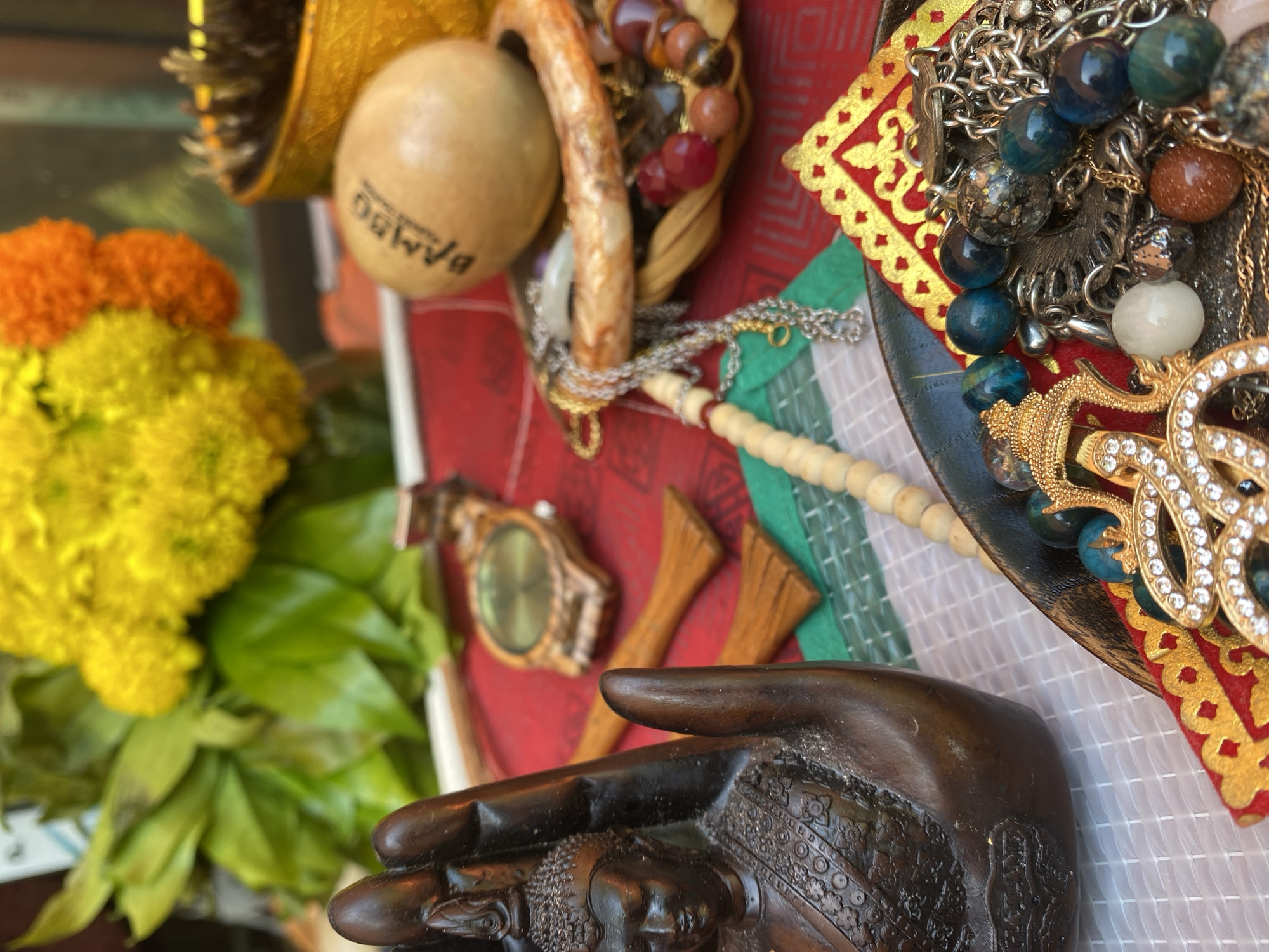 The image presents a close-up view of a tabletop assemblage of diverse objects, possibly related to spirituality or cultural practices. In the foreground, a dark, detailed hand sculpture, possibly depicting a Buddha figure, dominates the view. To the left, a bouquet of yellow and orange flowers with green leaves provides a vibrant contrast. On the red-toned table, a wooden watch and small wooden implements are visible. The background features a small woven basket containing beads, a wooden ball labeled "BAMBOO," and a plate adorned with jewelry including necklaces and beaded strands. The background also reveals decorative fabrics with red, gold, green, and white patterns. Given the presence of the Buddha hand and cultural elements, the image location is likely a home or shop in Baan Kai, Thailand.