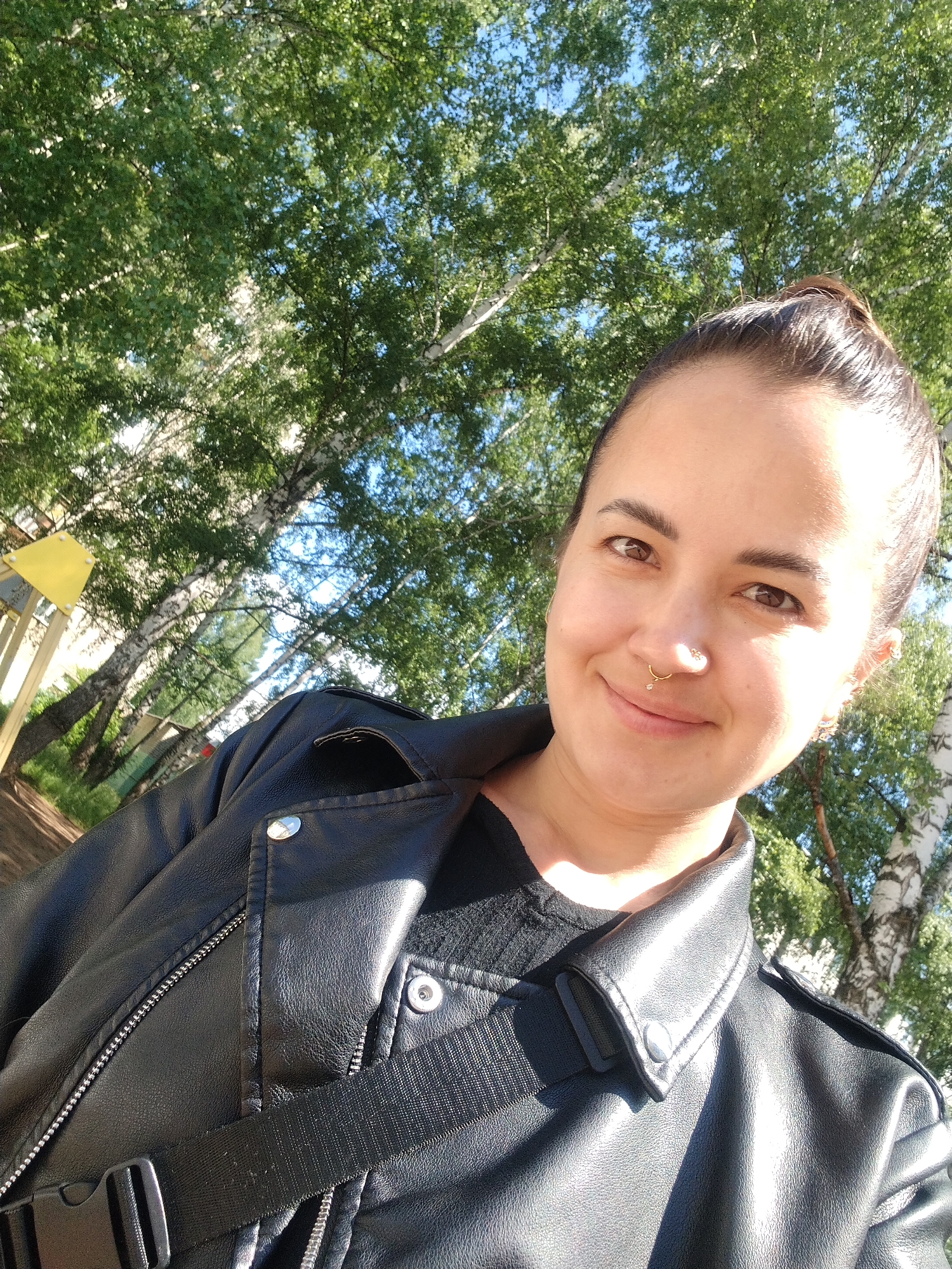 A brightly lit outdoor selfie captures a young adult woman with dark hair pulled back into a bun, smiling subtly at the camera. She has brown eyes, a gold septum piercing, and another small stud piercing on her nose, along with earrings. She is wearing a black leather jacket with silver hardware over a black top, and a black strap, possibly from a bag, crosses her chest.
The background is filled with the lush green canopy and distinctive white bark trunks of numerous birch trees, with sections of clear blue sky visible through the leaves. To the far left, a yellow structure, possibly part of playground equipment, is partially visible. The scene is illuminated by strong, direct sunlight, indicating a sunny day, likely morning or afternoon. The setting appears to be a park or natural outdoor area, consistent with the provided location of Chaikovsky, Russia. There is no visible text in the image.