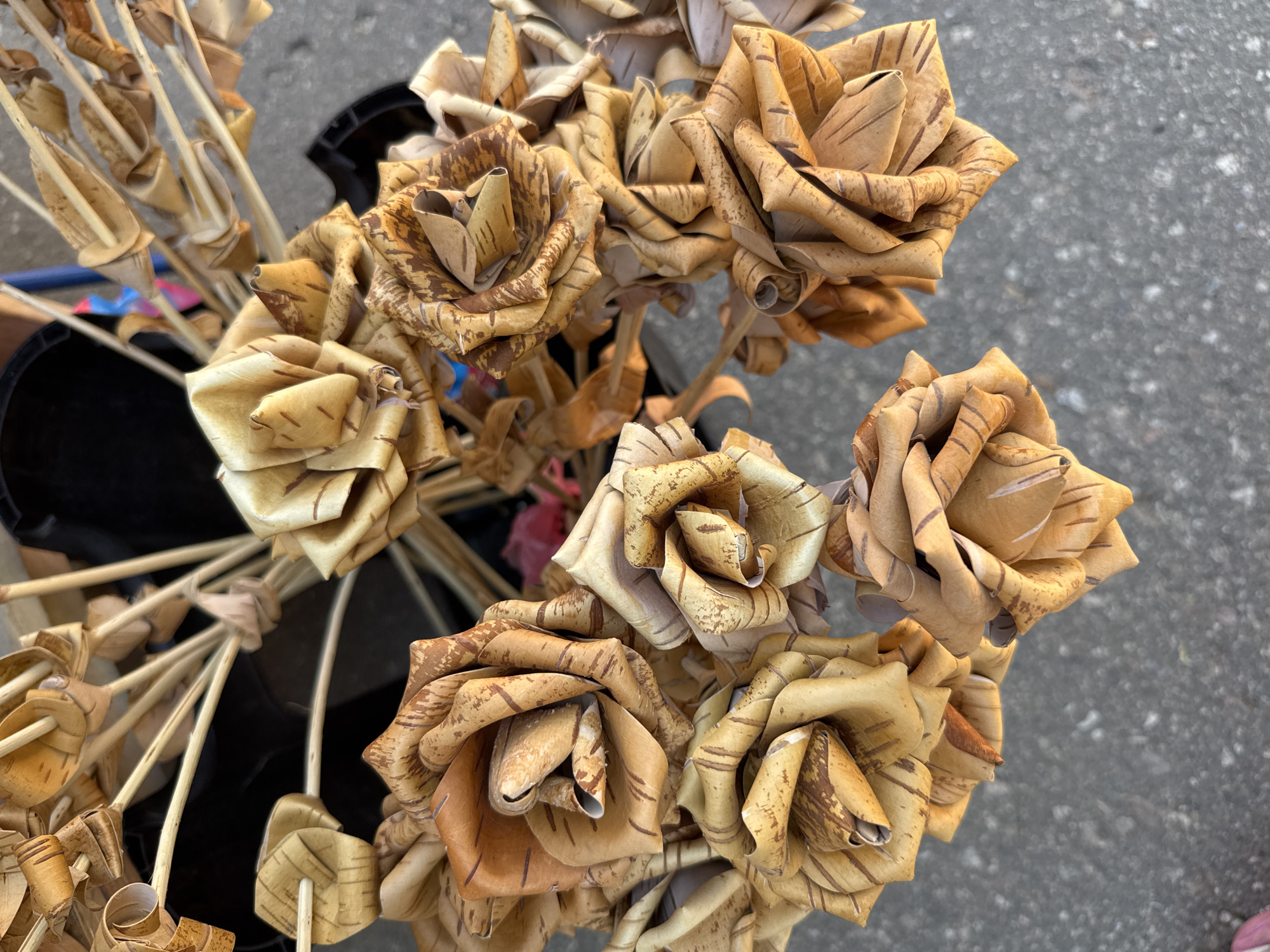 A bouquet of dried, artificial roses made from birch bark sits in a black container on a gray, asphalt surface. The roses are various shades of tan and brown, with natural bark markings visible on the petals. The stems are thin and light-colored. The background is blurred, but hints of a dark container and colorful objects, possibly other handicrafts, are visible. The scene appears to be outdoors, possibly at a market or street vendor stall. The image is taken from a top-down perspective. The location is Veliky Novgorod, Russia.