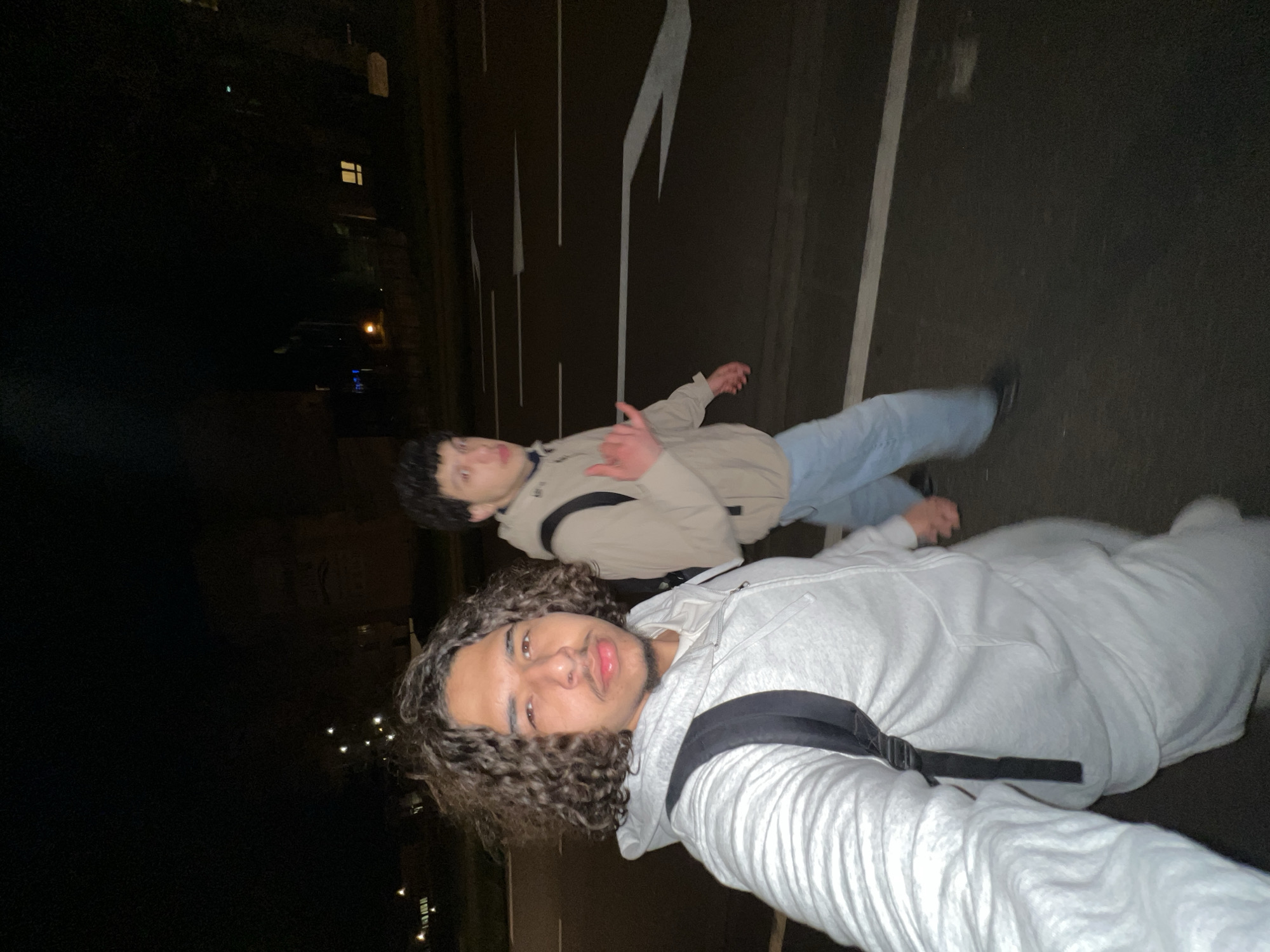 The image shows two young men standing on a road in Hoenheim, France at night. The man closest to the camera has curly hair and is wearing a light-colored hoodie with a black strap. He is looking at the camera. The other man is wearing a tan jacket, light blue jeans, and is holding up a hand gesture. They are standing on a road with white lane markings. In the background, there are buildings and street lights. The overall setting is urban and dimly lit.