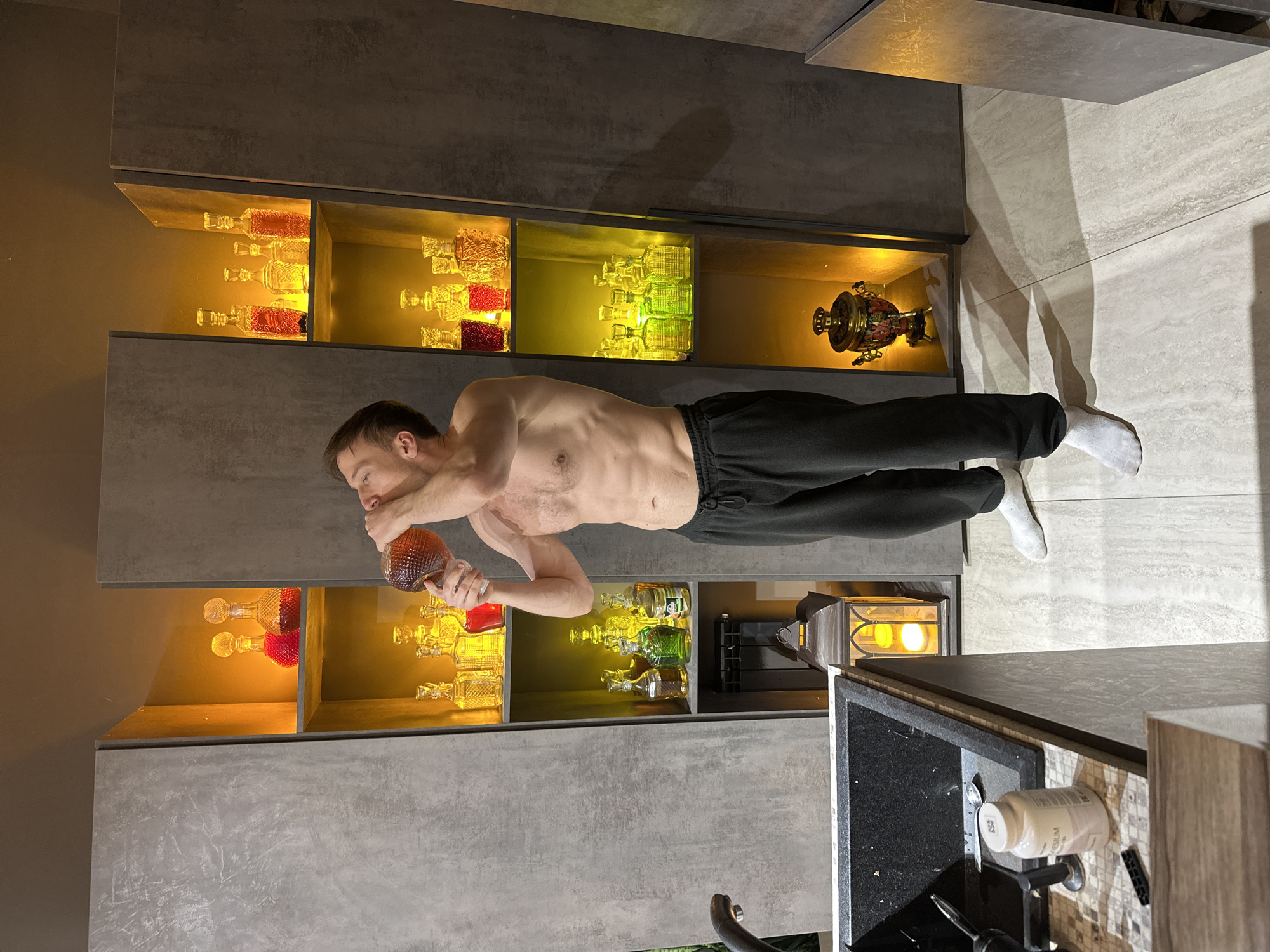 A shirtless man is drinking directly from a decorative glass bottle in a modern kitchen or dining area. He is wearing dark sweatpants and white socks. The backdrop consists of built-in gray cabinets with illuminated display shelves containing an assortment of decorative glass bottles and other objects including a samovar. The floor is tiled in a light gray pattern. A kitchen sink area with countertop items is visible on the right side of the frame. The photograph appears to be taken indoors with a focus on the man and the decorative displays. The location in Kursk, Russia, cannot be confirmed from the image.