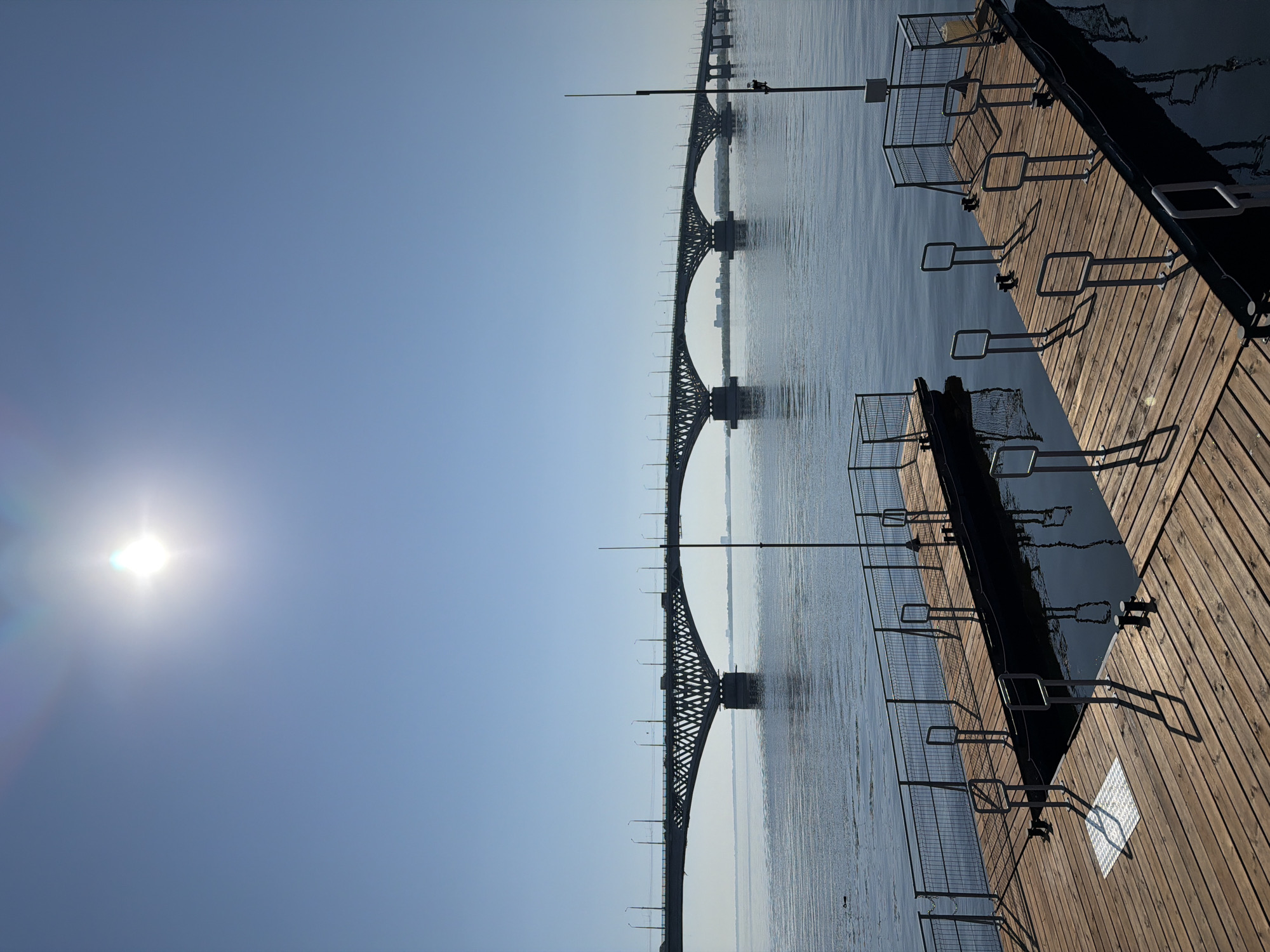 The image captures a wide view of a river with a long, arched bridge spanning across it. In the foreground, two wooden piers with metal railings and upright handrails extend into the calm water. The sun is bright and high in the clear blue sky, casting shadows on the piers. The bridge appears to be an elevated roadway with vehicles visible on it. In the distance, on the far bank of the river, a city skyline with buildings can be seen. The overall scene suggests a sunny day in an urban environment by a river. This is likely the Saratov Bridge, a prominent landmark connecting the cities of Saratov and Engels across the Volga River in Saratov Oblast, Russia.
