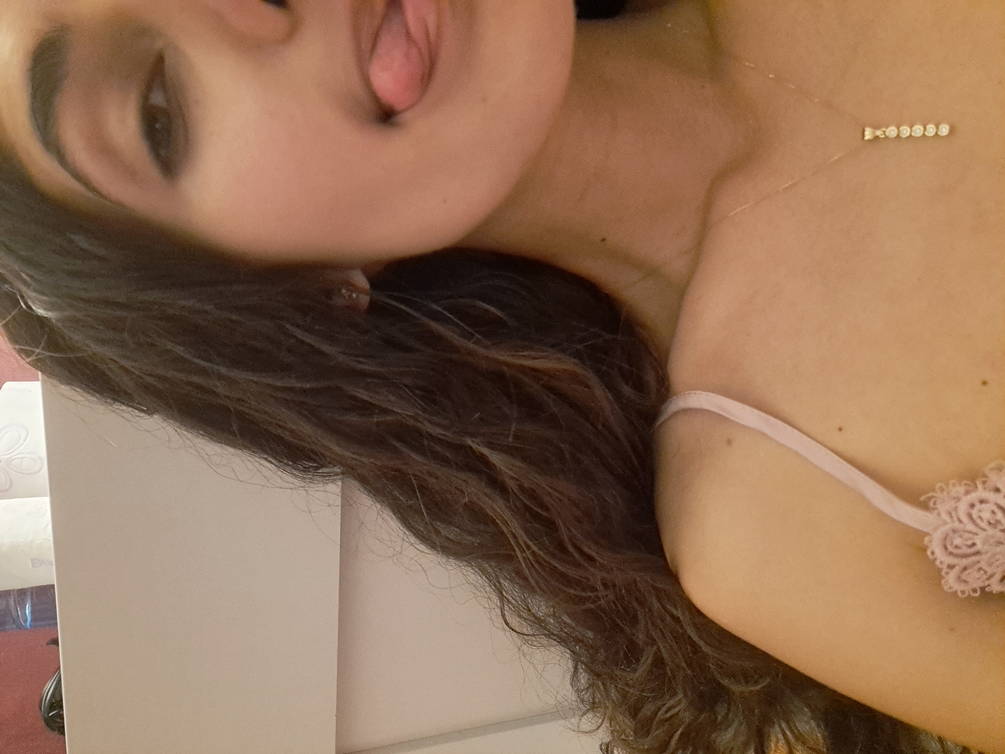 In a close-up, over-the-shoulder shot, a young woman with dark, curly hair is pictured. Her right shoulder and neck are visible, and she is wearing a light pink camisole with delicate lace detailing on the strap. A delicate gold necklace with a pendant of small, circular stones hangs around her neck. Her face is partially visible, with her right eye looking towards the camera. Her tongue is sticking out to the right, and her lips are slightly parted. Her skin tone is fair. The background is indistinct, with hints of what appears to be a white surface and some partially visible items that may be toiletries. The lighting appears to be indoor and soft. The image is taken from an eye-level perspective.