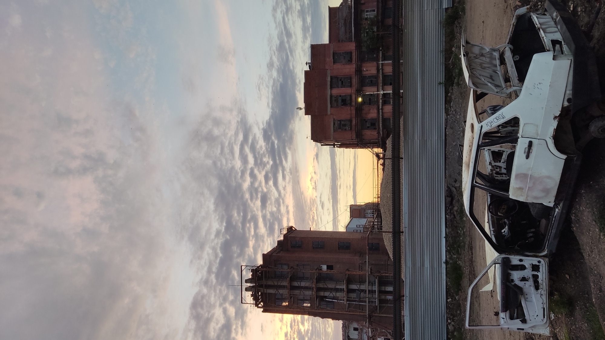 This media file captures a post-apocalyptic industrial scene at dusk in Krasnodar, Russia. The primary subjects are two dilapidated brick buildings and a gutted white car. The buildings, appearing abandoned and in disrepair, feature broken windows and skeletal scaffolding, hinting at a past industrial function. The car, stripped of its interior and exterior panels, lies on its side, exposing its skeletal frame.
The scene is bathed in the soft, warm light of a setting sun, casting long shadows and creating a dramatic sky filled with scattered clouds. The time of day is clearly dusk, with the golden hues of the sunset transitioning into twilight. The overall mood is one of decay, abandonment, and desolation. The location cues, such as the industrial architecture and the specific type of vehicle, along with the provided context of Krasnodar, Russia, firmly place the scene in an urban industrial periphery.
There is visible text on the side of the white car, partially obscured. It appears to be a graffiti tag or inscription, but it is too blurry to be fully deciphered. The activity taking place is none; the image freezes a moment in time, focusing on the static state of decay. The lack of human presence reinforces the feeling of abandonment and stillness.