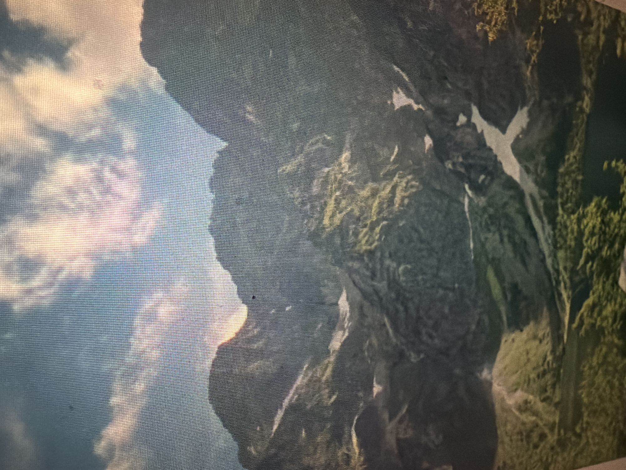 The image displays a panoramic view of a mountainous landscape under a partly cloudy sky. The mountains are rugged and covered in patches of green vegetation and snow. Sunlight filters through the clouds, casting a warm glow on the scene and highlighting the textures of the mountains and the sky. The lower portion of the image shows a valley with some trees and a body of water, possibly a lake. The scene is serene and captures the grandeur of nature. This image is not located in the city of Baan Mae Nam, Thailand.