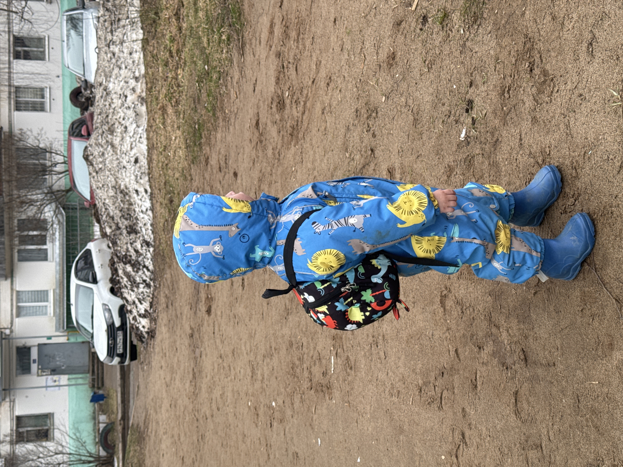 A child, dressed in a blue, animal-themed waterproof jumpsuit and matching blue rain boots, stands on a sandy patch of ground. The child has a small, black, printed bag slung across their body. The background includes a snowy area, bare trees, several parked cars (including a white van), and a multi-story building with visible windows. The location is outdoors, and based on the description, it can be identified as an area in the city of Chaikovsky, Russia.