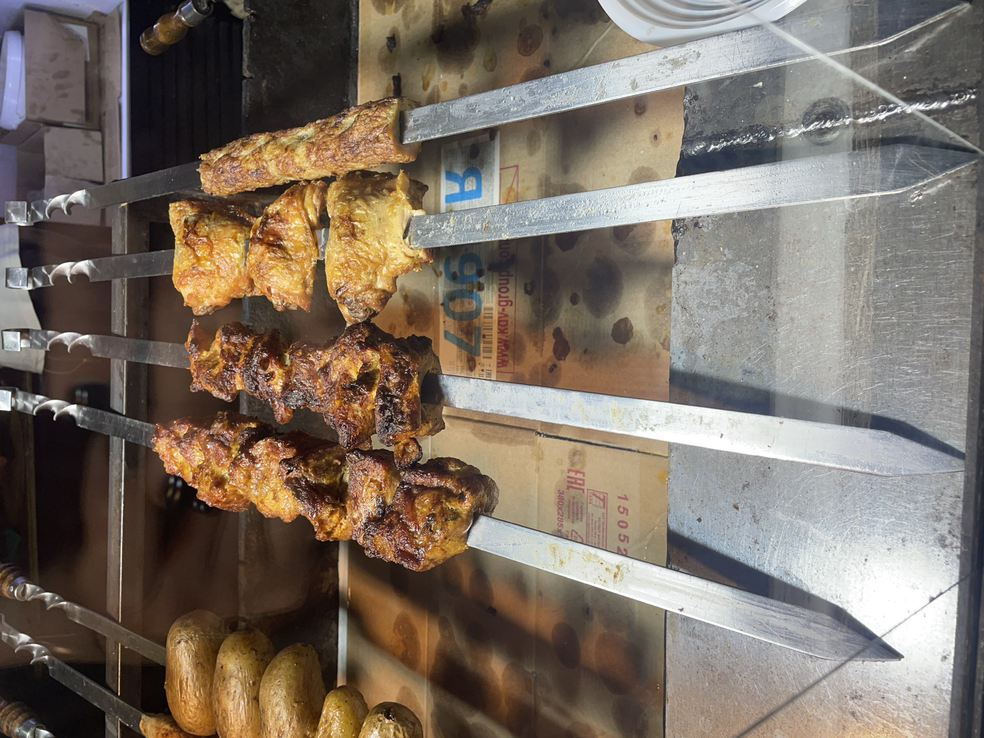 The image shows skewers of grilled meat and potatoes at a food stand. There are at least three skewers visible, each holding several pieces of browned meat. To the left, there are also several whole potatoes on skewers. The food is displayed over what appears to be a cardboard surface with some printing on it, including what looks like part of a website address and some numbers. In the background, there are metal shelves and what appear to be some white boxes. The overall impression is of food being prepared for sale. The scene is not specific to any particular city or location.