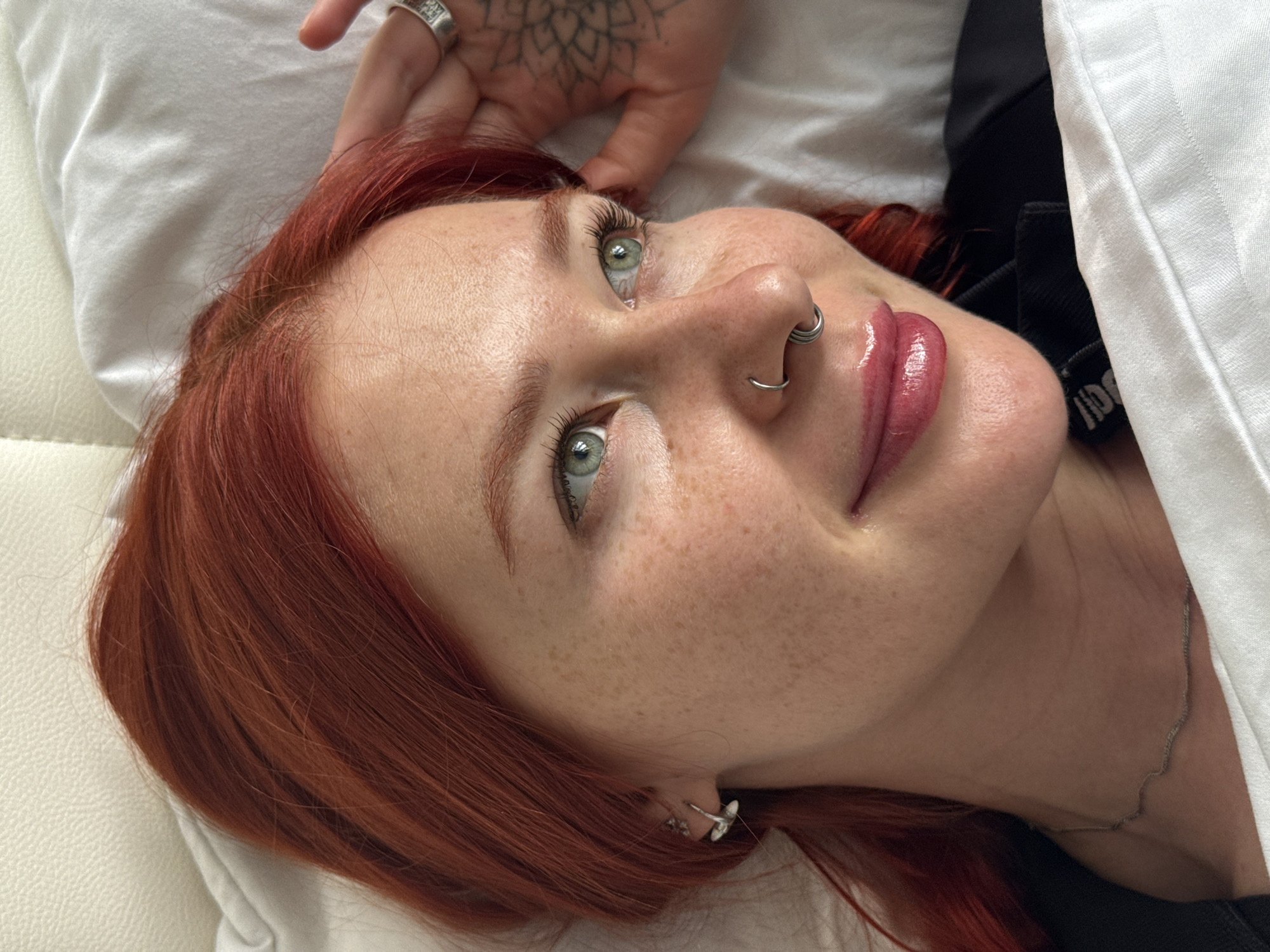 A close-up shot features a fair-skinned person with red hair lying on a white cushion. The person's face is slightly angled, and they have freckles across their face. Their eyes are a light green color. The person has a nose ring and is wearing earrings. One hand with a tattoo on it is raised slightly near the head. A necklace can be seen around their neck. The location in Rostov-on-Don, Russia is not discernible from the image.