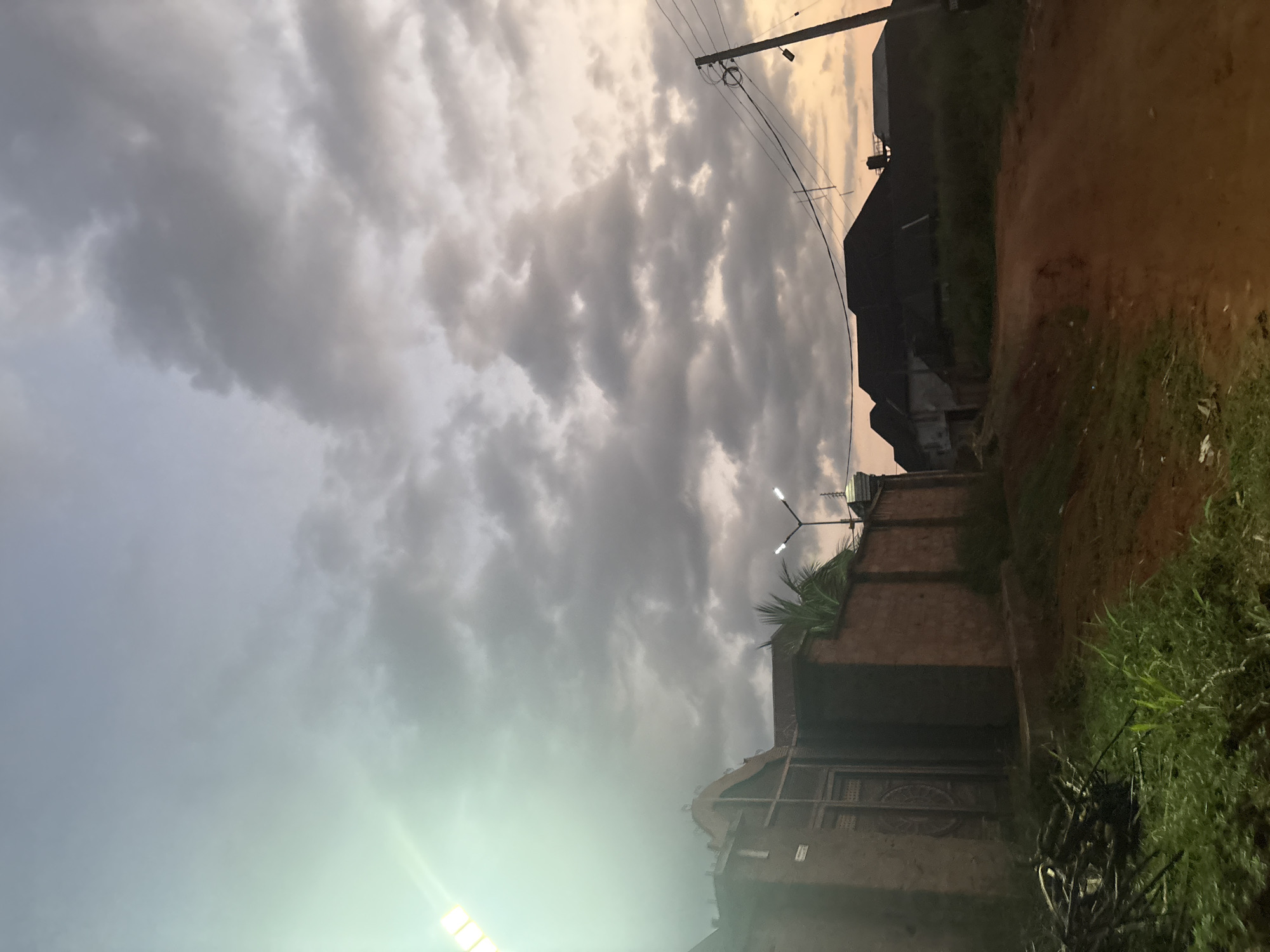 The image depicts a street scene in Obosi, Nigeria, likely during the early morning or late evening due to the soft, diffused light and cloudy sky. The foreground features a dirt road, typical of many areas in Nigeria, with sparse green vegetation and some fallen branches on the left side.
On the left side of the road, a partially visible building constructed from reddish-brown bricks is prominent. It has an ornate metal gate, suggesting a residential property or a gated community. Above this building, a bright, artificial light source, possibly a streetlamp or a security light, casts a strong green glow and lens flare across the upper left portion of the image.
Further down the road, to the right, are silhouetted structures of houses with dark, sloped roofs. Overhead, power lines crisscross the sky, supported by utility poles. The sky itself is filled with a mix of grey and white clouds, with a subtle warm glow on the right horizon, indicating either sunrise or sunset. The overall atmosphere is somewhat overcast, with the lighting creating a muted, subdued tone across the scene. No people are visible in the frame.