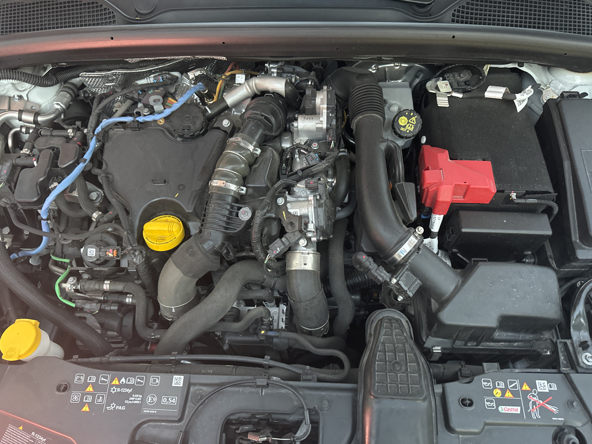 The image shows the engine bay of a car, viewed from above. The engine is complex, with many hoses, wires, and metal components. There are also several plastic containers and covers. A red component, possibly a fuse box or battery terminal cover, is visible in the lower right portion of the image. The engine bay appears to be clean and well-maintained. The background is blurred, but suggests an outdoor environment. There are no people or animals visible in the image. The location of Murcia, Spain is not explicitly indicated by any visual cues in the photograph.