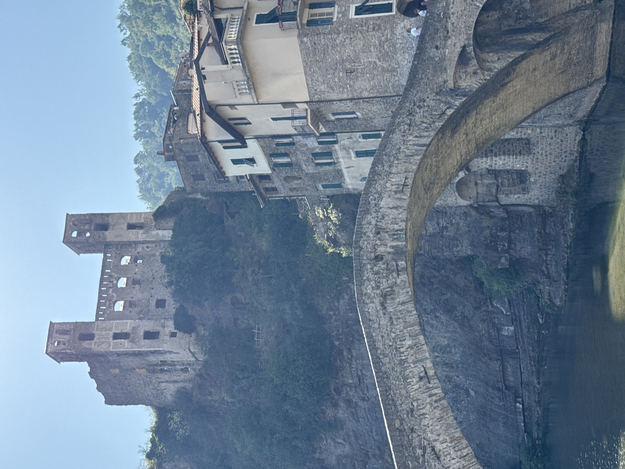 A medieval stone bridge with a single arch spans a calm body of water. On the far bank, buildings made of stone and plaster are nestled into the hillside, with terracotta tiled roofs. To the left and higher up the hillside, a ruined castle stands against a clear blue sky, with lush green trees surrounding it. The castle has two prominent towers and remnants of walls with arched windows. Several people are visible on the bridge and in front of the buildings. The overall scene depicts a historic European town.